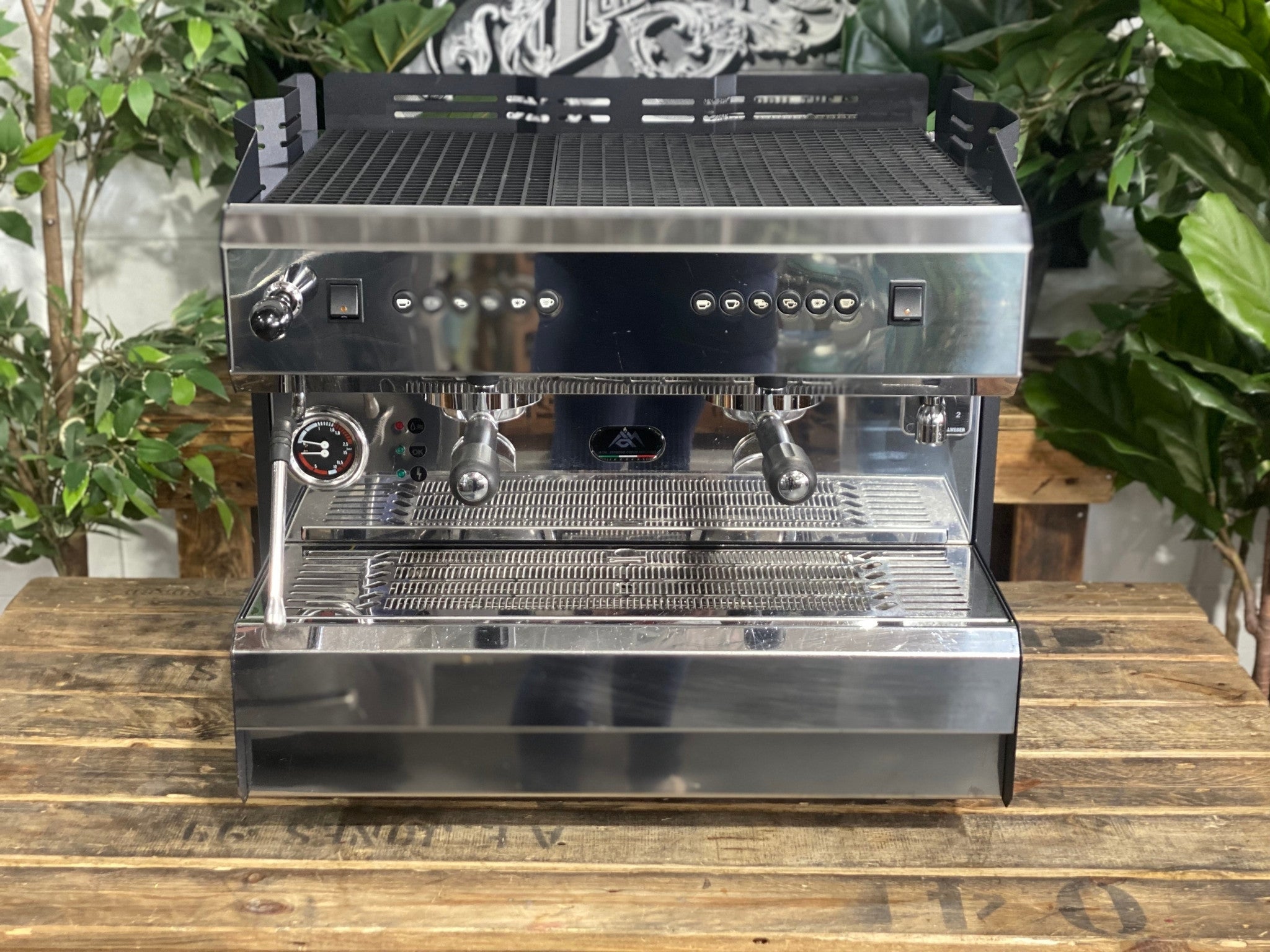 ACM Teknica 2 Group Black Espresso Coffee Machine sitting on a wooden bench. There is greenery in the background. The image is taken from the front of the machine but a higher angle so that you can see the front of the machine and also the top of the machine