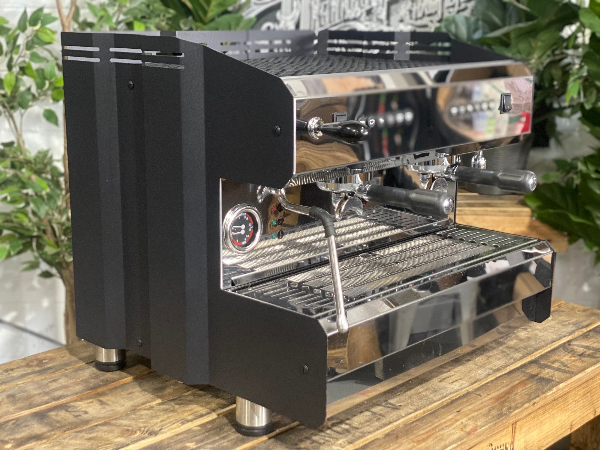 ACM Teknica 2 Group Black Coffee Machine sitting on a wooden bench. There is greenery in the background. The espresso machine is on an angle facing the right.