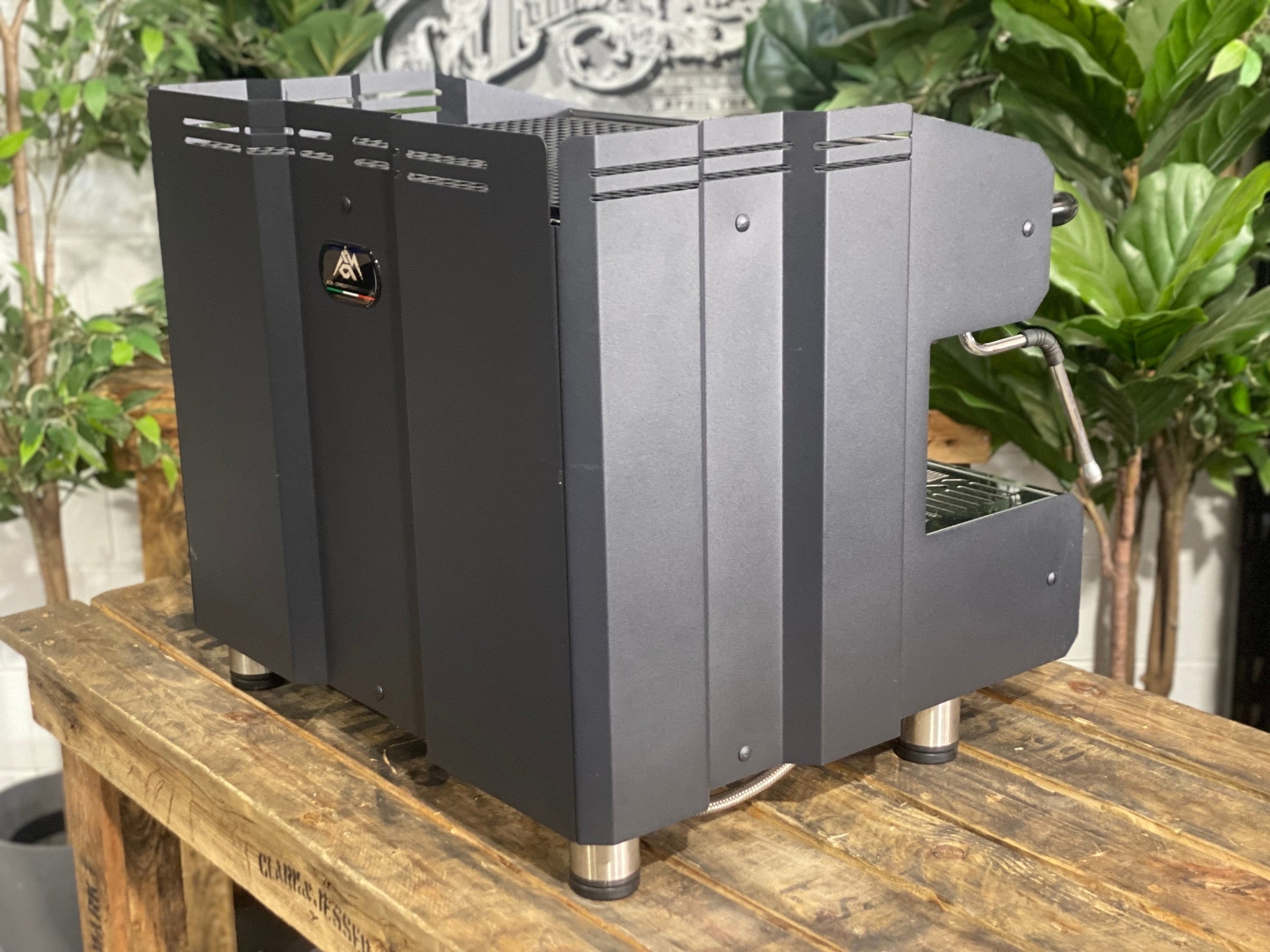 ACM Teknica 2 Group Black Coffee Machine sitting on a wooden bench. There is greenery in the background. The image is taken from behind the machine and the machine is sitting on an angle so that you can see the back of the machine and the side