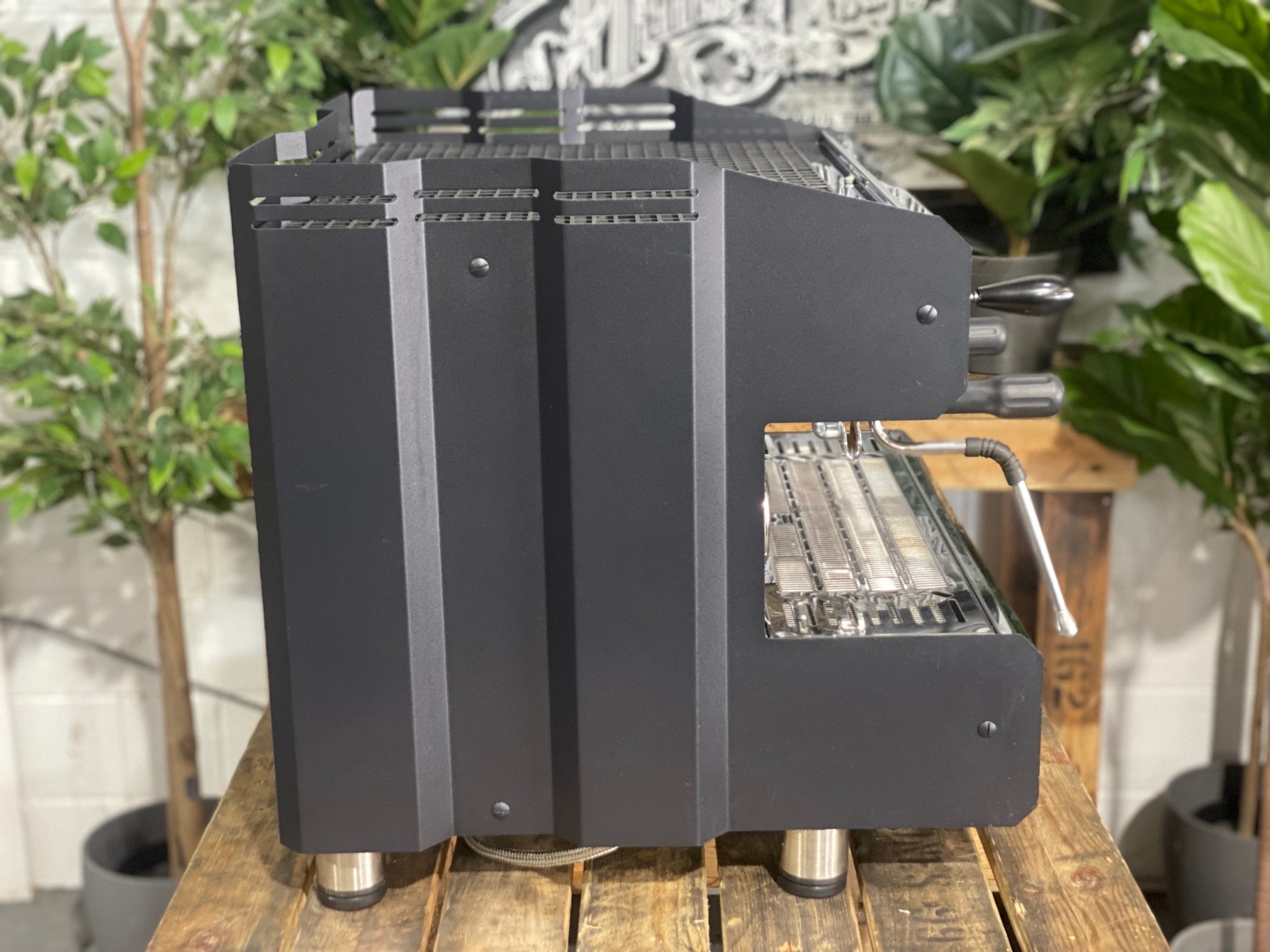 ACM Teknica 2 Group Black Espresso Coffee Machine sitting on a wooden bench. There is greenery in the background. The image is taken from the side of the machine.