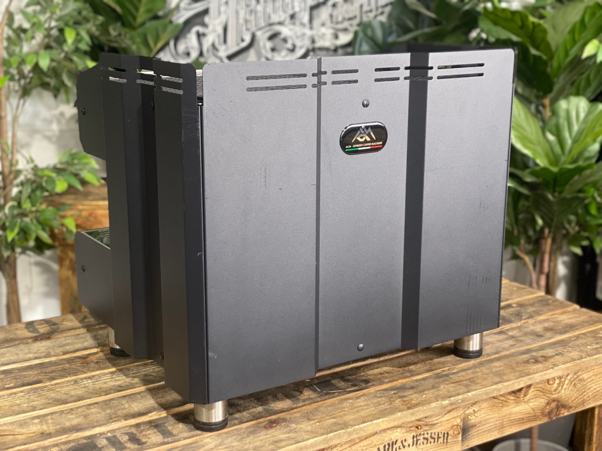 ACM Teknica 2 Group Black Espresso Coffee Machine sitting on a wooden bench. There is greenery in the background. The image is taken from behind the machine and the machine is sitting on an angle so that you can see the back of the machine and part of the the side