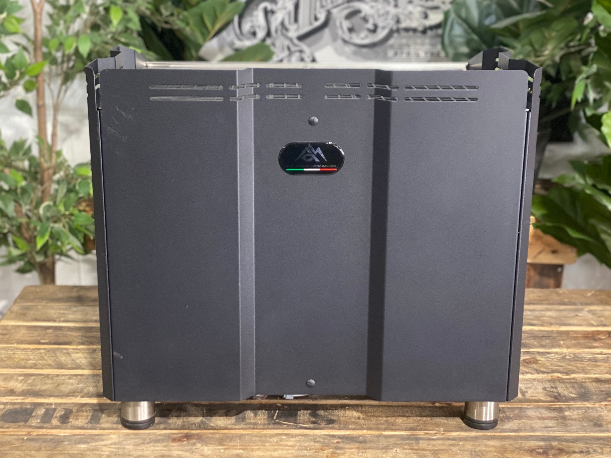 ACM Teknica 2 Group Black Espresso Coffee Machine sitting on a wooden bench. There is greenery in the background. The image is taken from behind the machine so the back is visible.