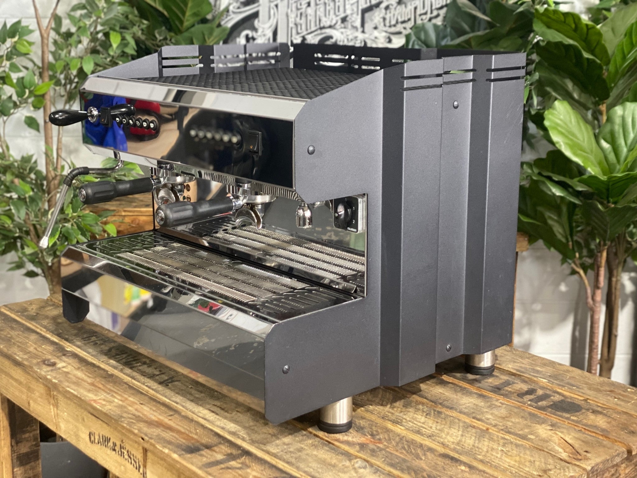 ACM Teknica 2 Group Black Espresso Coffee Machine sitting on a wooden bench. There is greenery in the background. The image is taken from the side of the machine and the machine is sitting on an angle so that you can see the front of the machine and the side