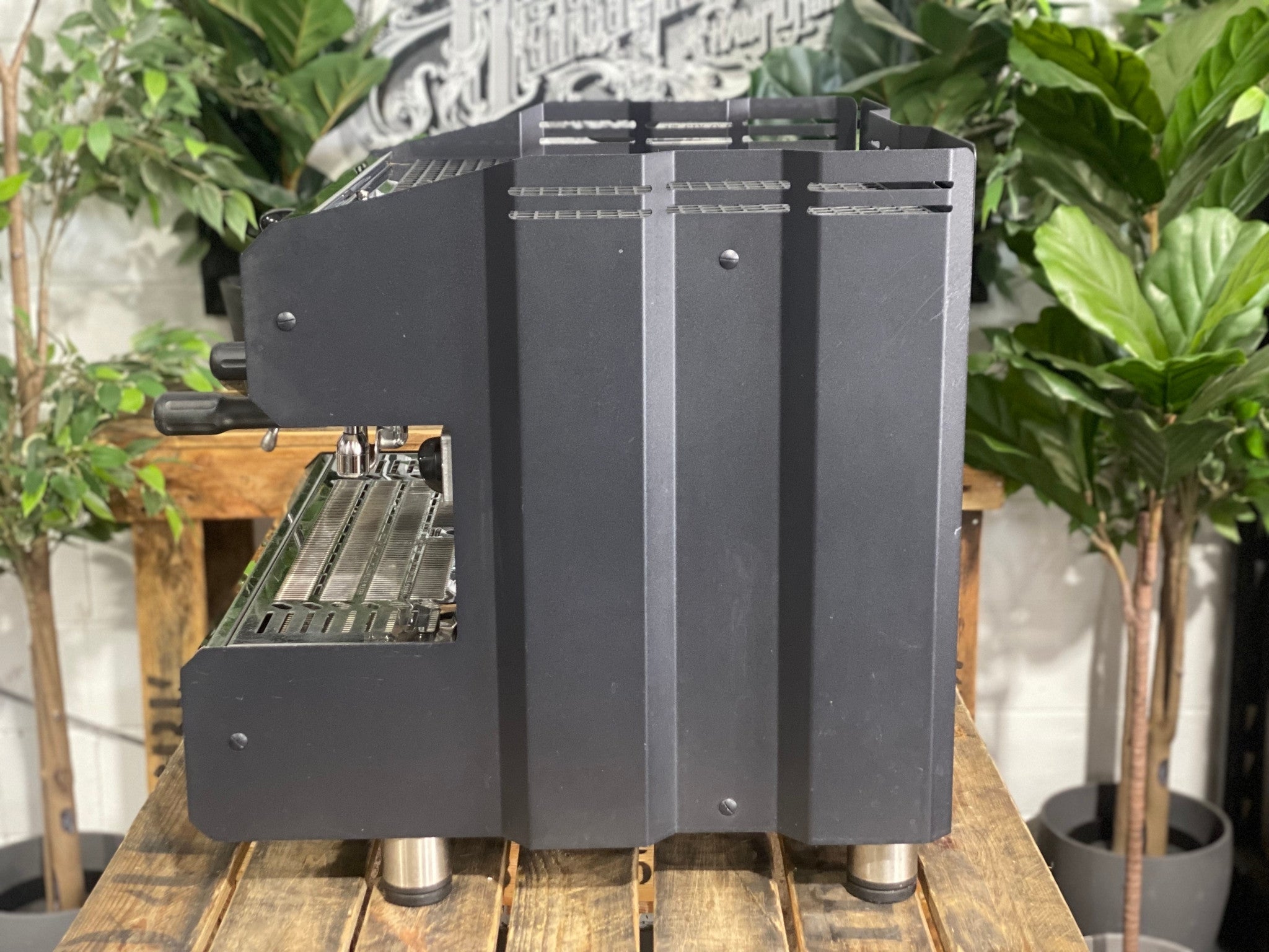 ACM Teknica 2 Group Black Espresso Coffee Machine sitting on a wooden bench. There is greenery in the background. The image is taken from the side of the machine