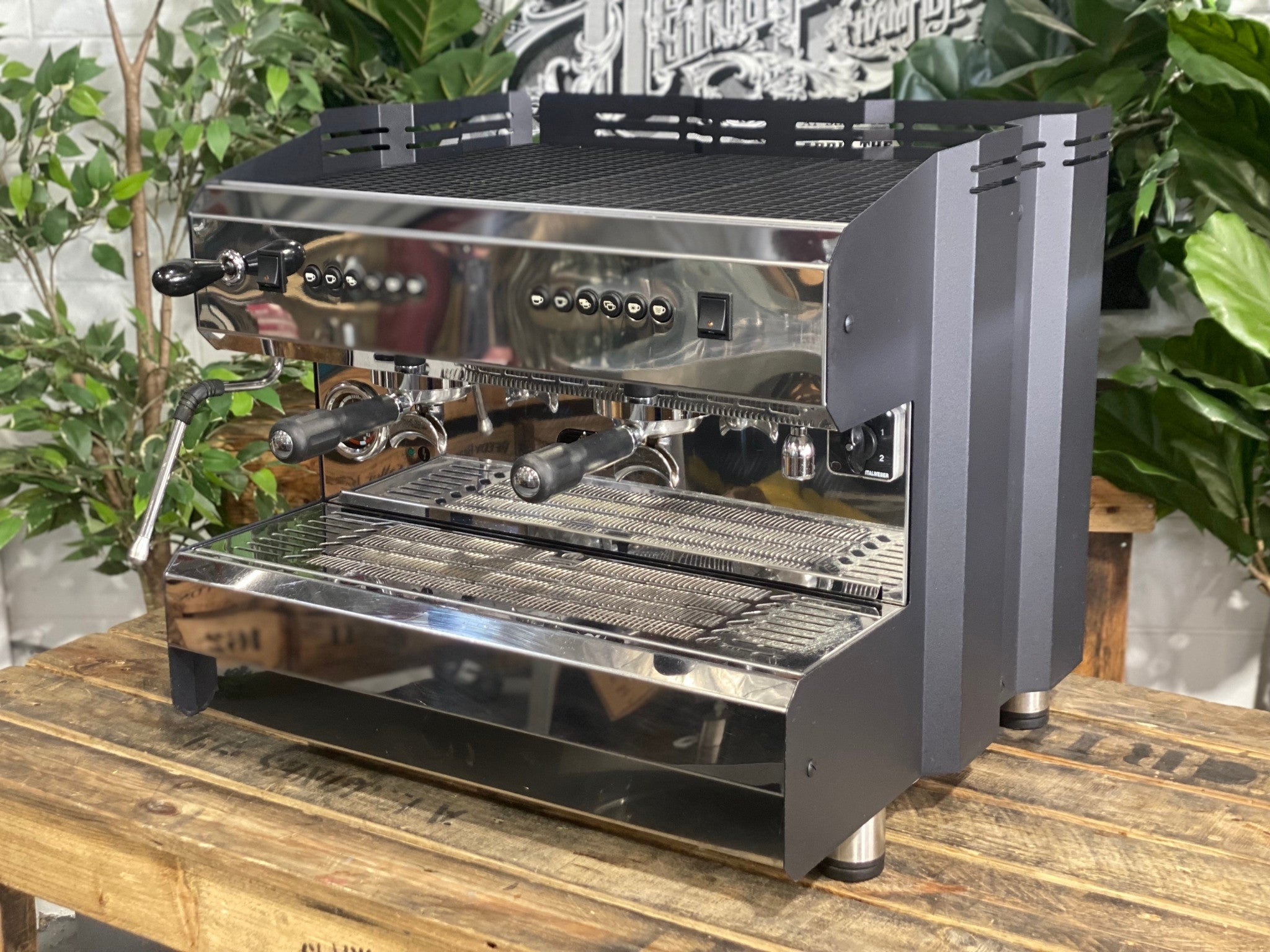 ACM Teknica 2 Group Black Espresso Coffee Machine sitting on a wooden bench. There is greenery in the background. The image is taken from the front of the machine and the machine is sitting on an angle so that you can see the front of the machine and the side