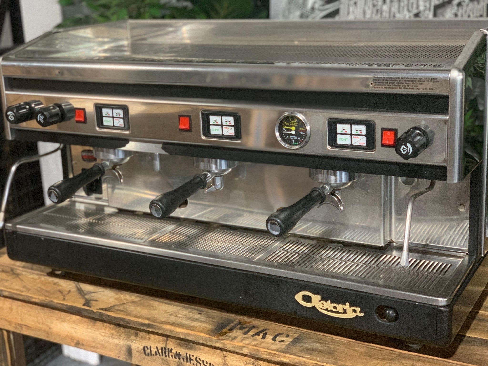 Astoria Argenta 3 Group Espresso Coffee Machine sitting on a wooden bench, there is greenery in the background. The machine is sitting on an angle facing slightly left and the image is a close up image of the front dials of the machine