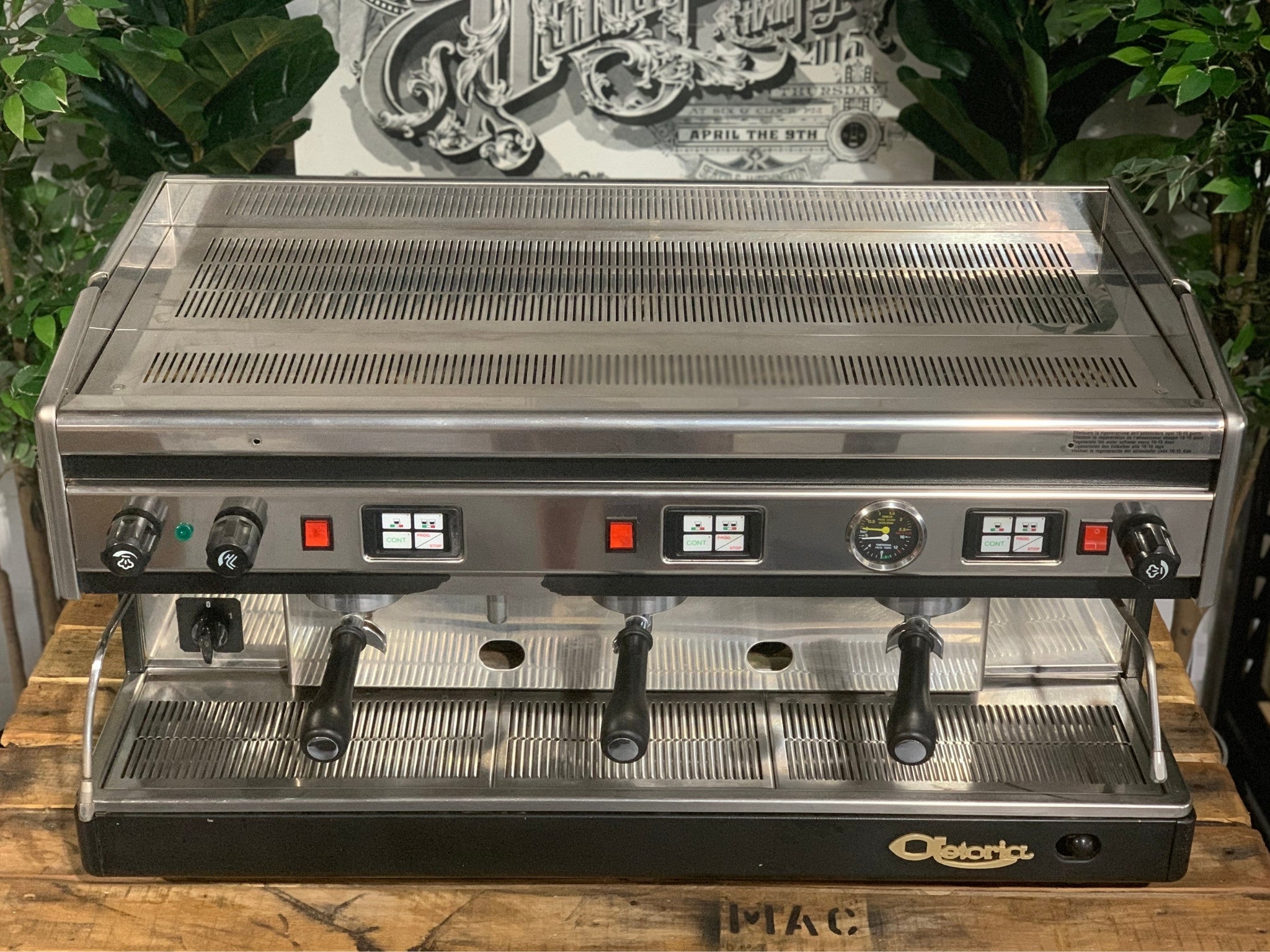 Astoria Argenta 3 Group Espresso Coffee Machine sitting on a wooden bench, there is greenery in the background. The image is taken from the front of the machine from a high angle so the top of the machine is visible.