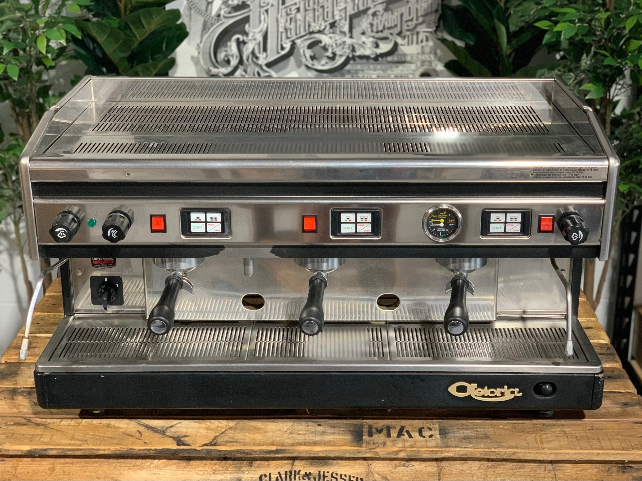 Astoria Argenta 3 Group Espresso Coffee Machine sitting on a wooden bench, there is greenery in the background. The image is taken from the front of the machine and the top of the machine is also visible.