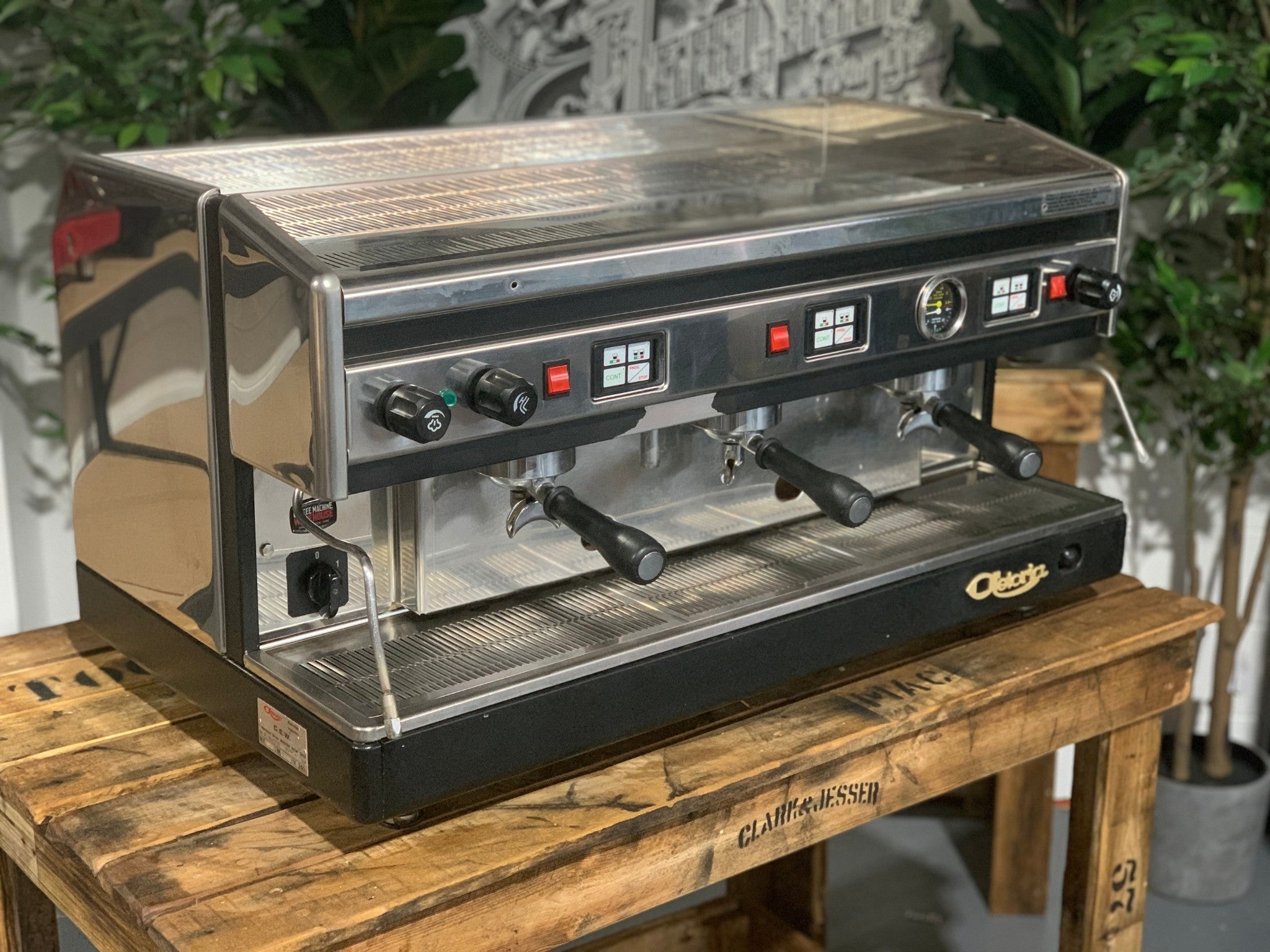 Astoria Argenta 3 Group Espresso Coffee Machine sitting on a wooden bench, there is greenery in the background. The image is taken from the side corner of the machine and the machine is on an angle facing right so the front and side are visible,