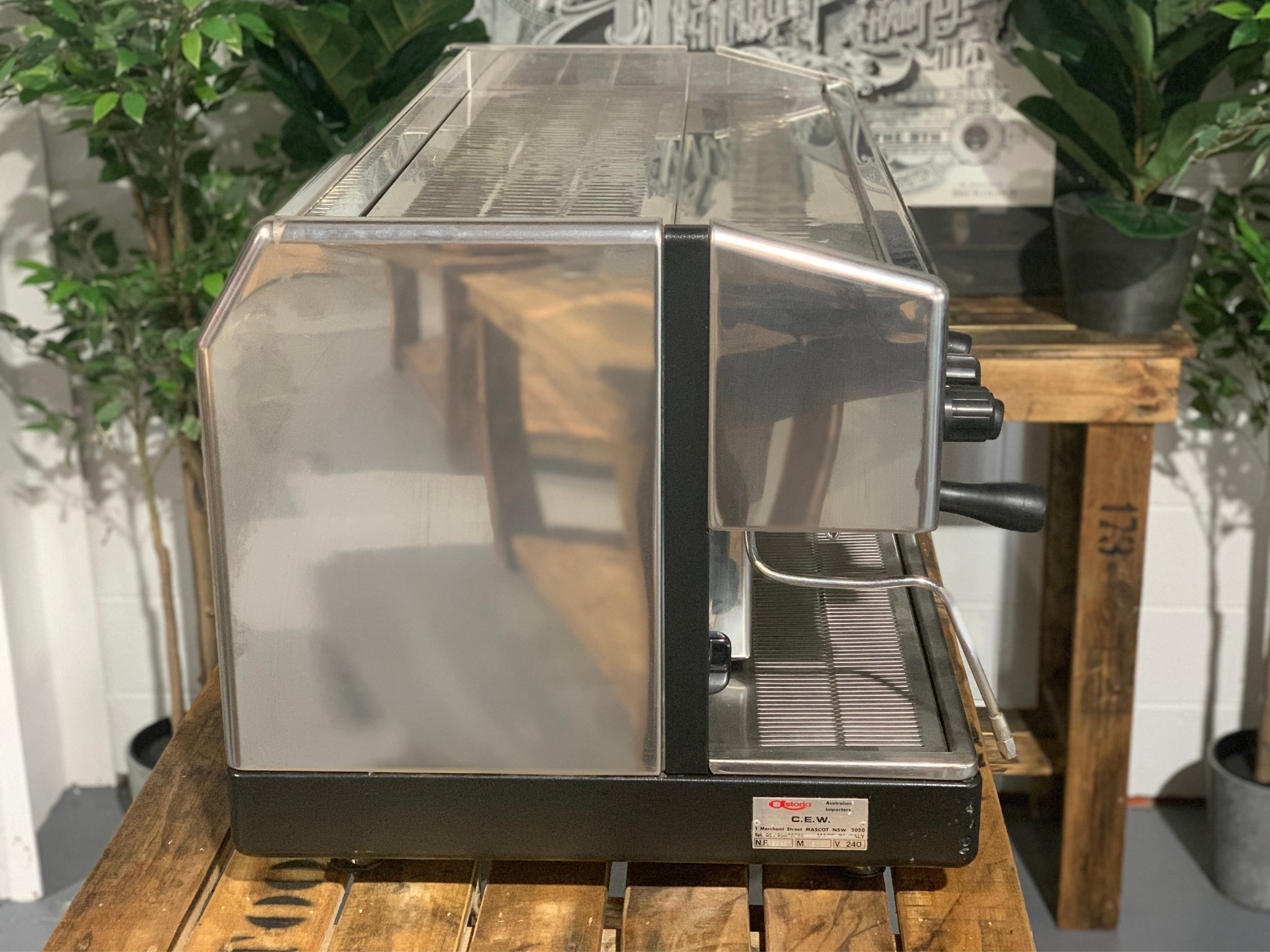 Astoria Argenta 3 Group Espresso Coffee Machine sitting on a wooden bench, there is greenery in the background. The image is taken from the side of the machine