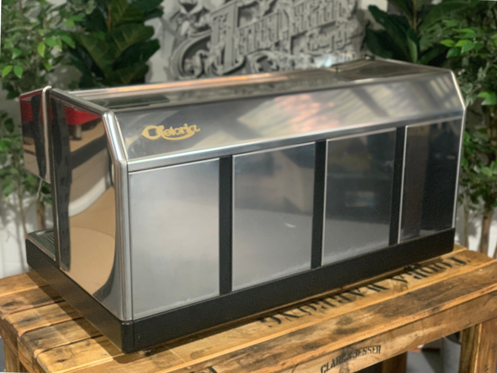 Astoria Argenta 3 Group Espresso Coffee Machine sitting on a wooden bench, there is greenery in the background. The image is taken from the back of the machine and the machine is on an angle facing left making part of the side visible as well as the back of the machine .