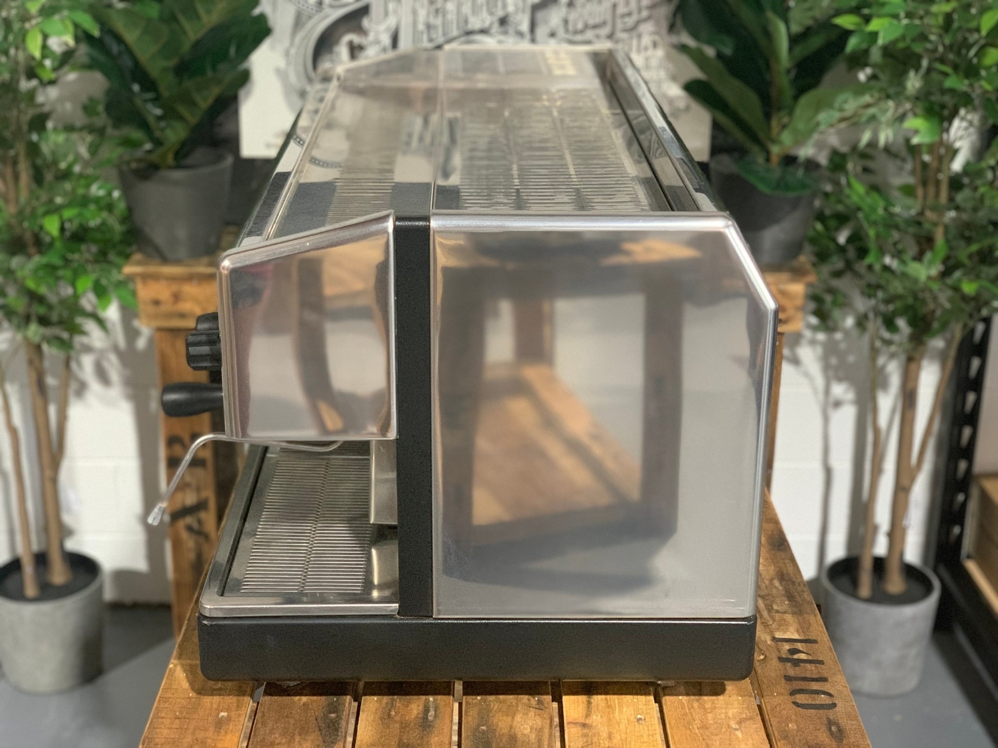 Astoria Argenta 3 Group Espresso Coffee Machine sitting on a wooden bench, there is greenery in the background. The image is taken from the side of the machine.