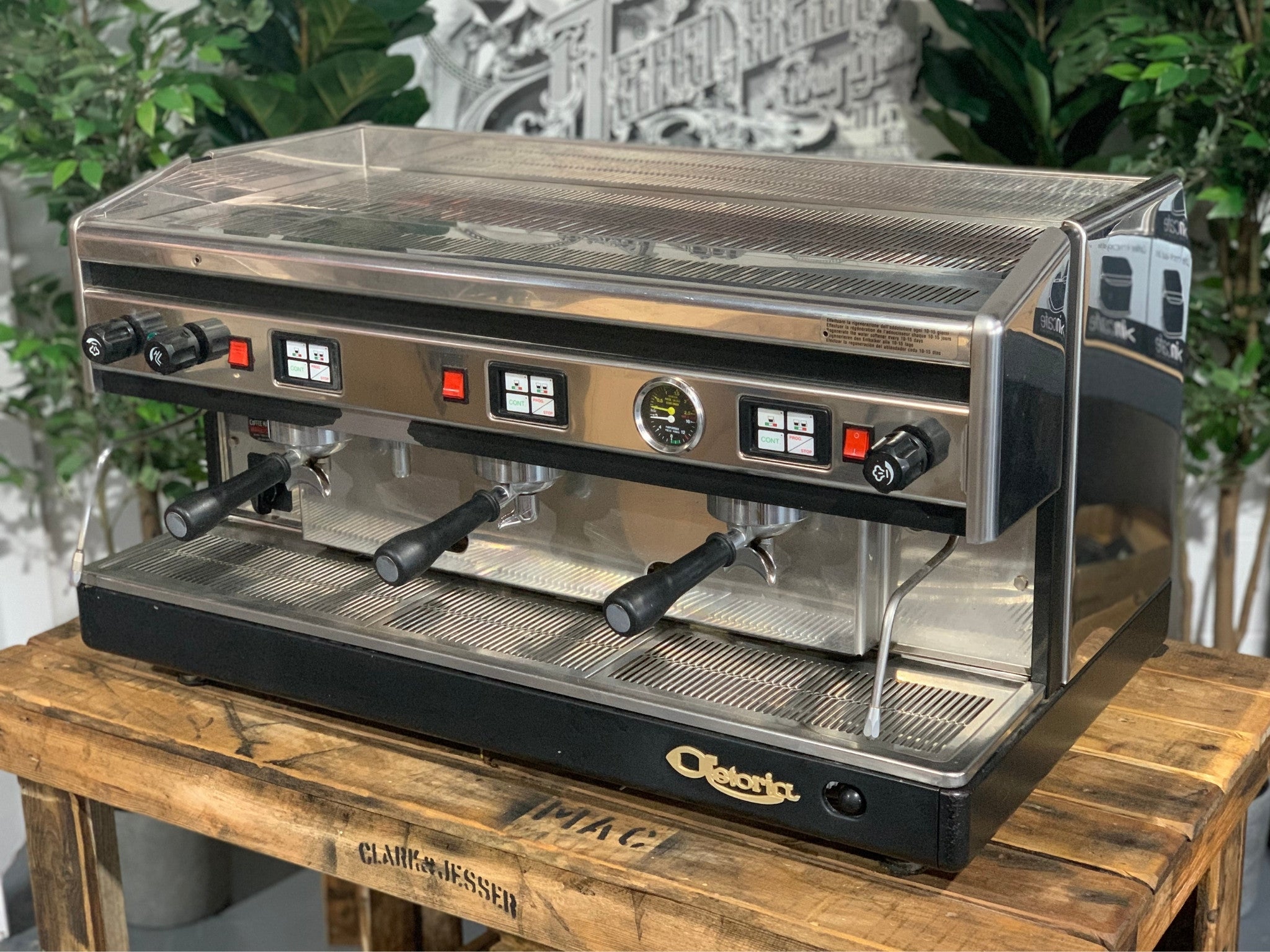 Astoria Argenta 3 Group Espresso Coffee Machine sitting on a wooden bench, there is greenery in the background. The machine is sitting on an angle facing left so you can see the front of the machine and part of the side.