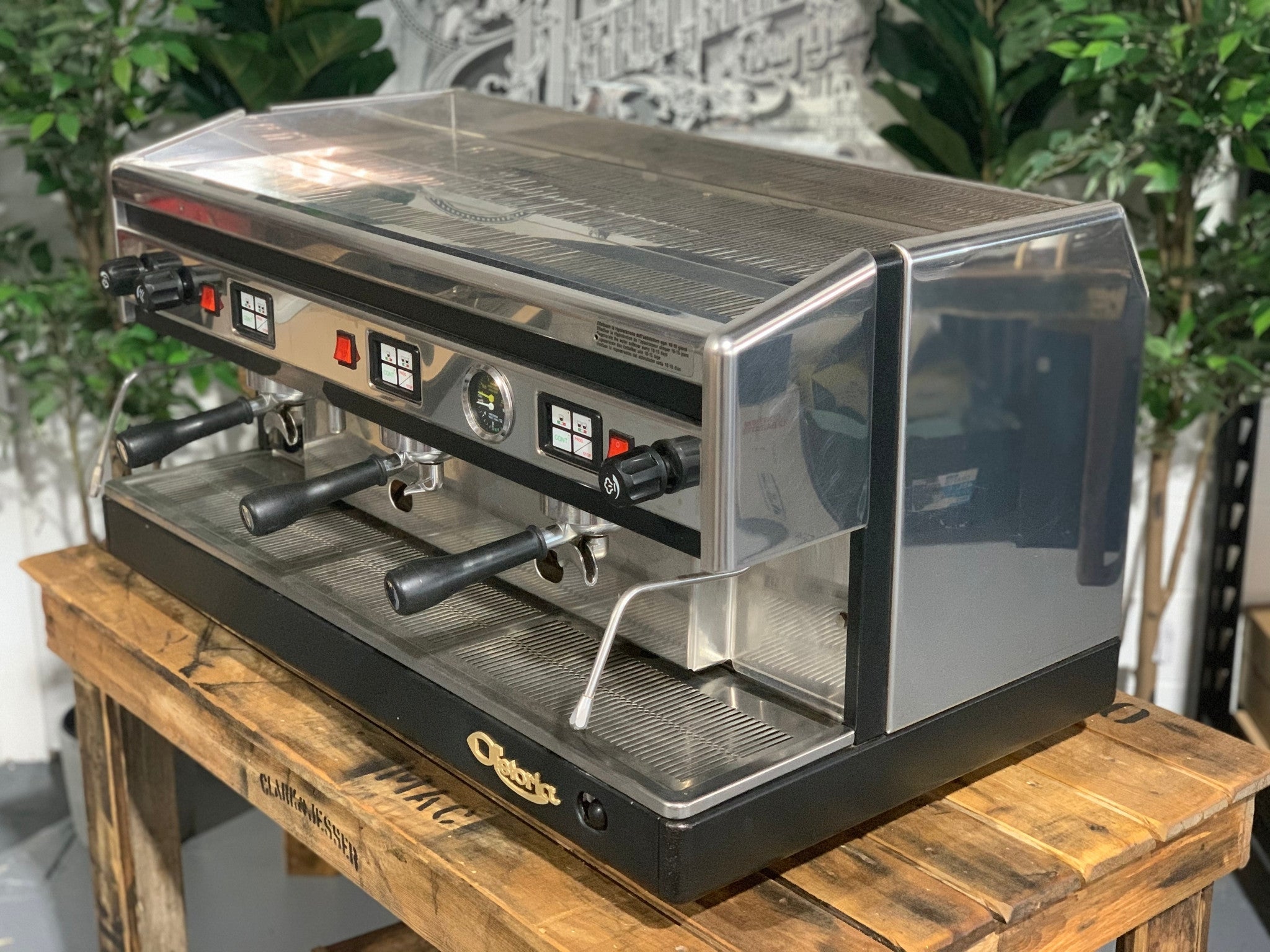 Astoria Argenta 3 Group Espresso Coffee Machine sitting on a wooden bench, there is greenery in the background. The machine is sitting on an angle facing left so you can see the front of the machine and also the side.