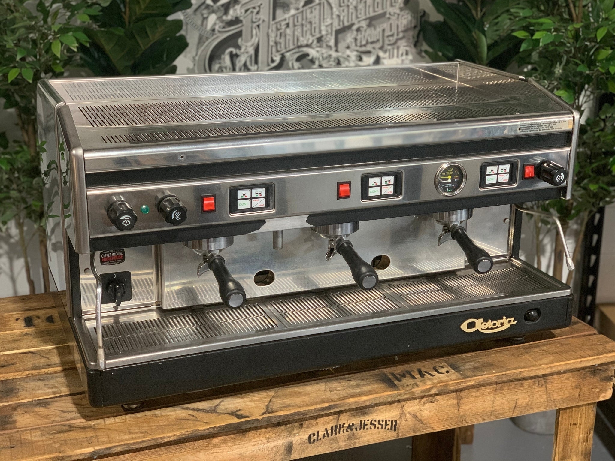 Astoria Argenta 3 Group Espresso Coffee Machine sitting on a wooden bench, there is greenery in the background. The image is taken from the front of the machine and the machine is on an angle facing right making part of the side visible .