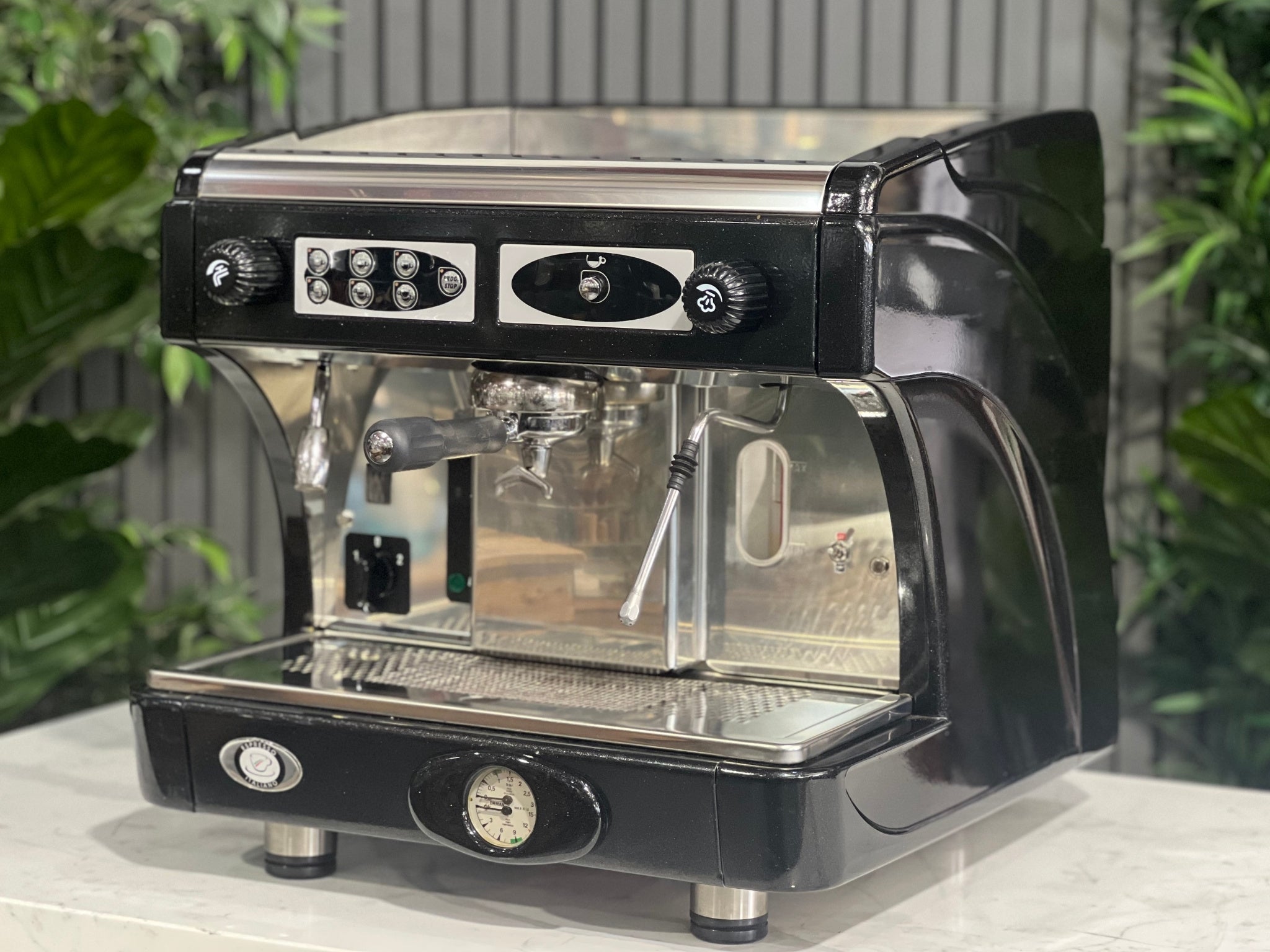 Astoria Calypso 1 Group Black Espresso Coffee Machine sitting on a marble bench with greenery in the background. Image is taken from an angle so that the front and side of machine is visible.