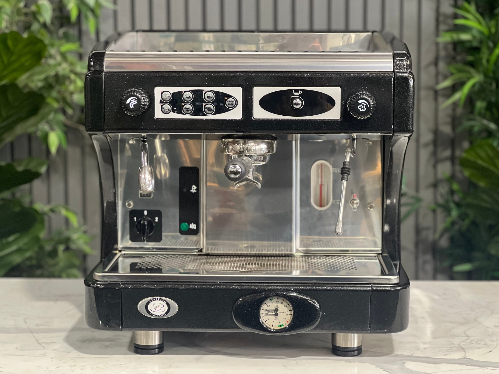Astoria Calypso 1 Group Black Espresso Coffee Machine sitting on a marble bench with greenery in the background.  Image is taken from the front of the machine