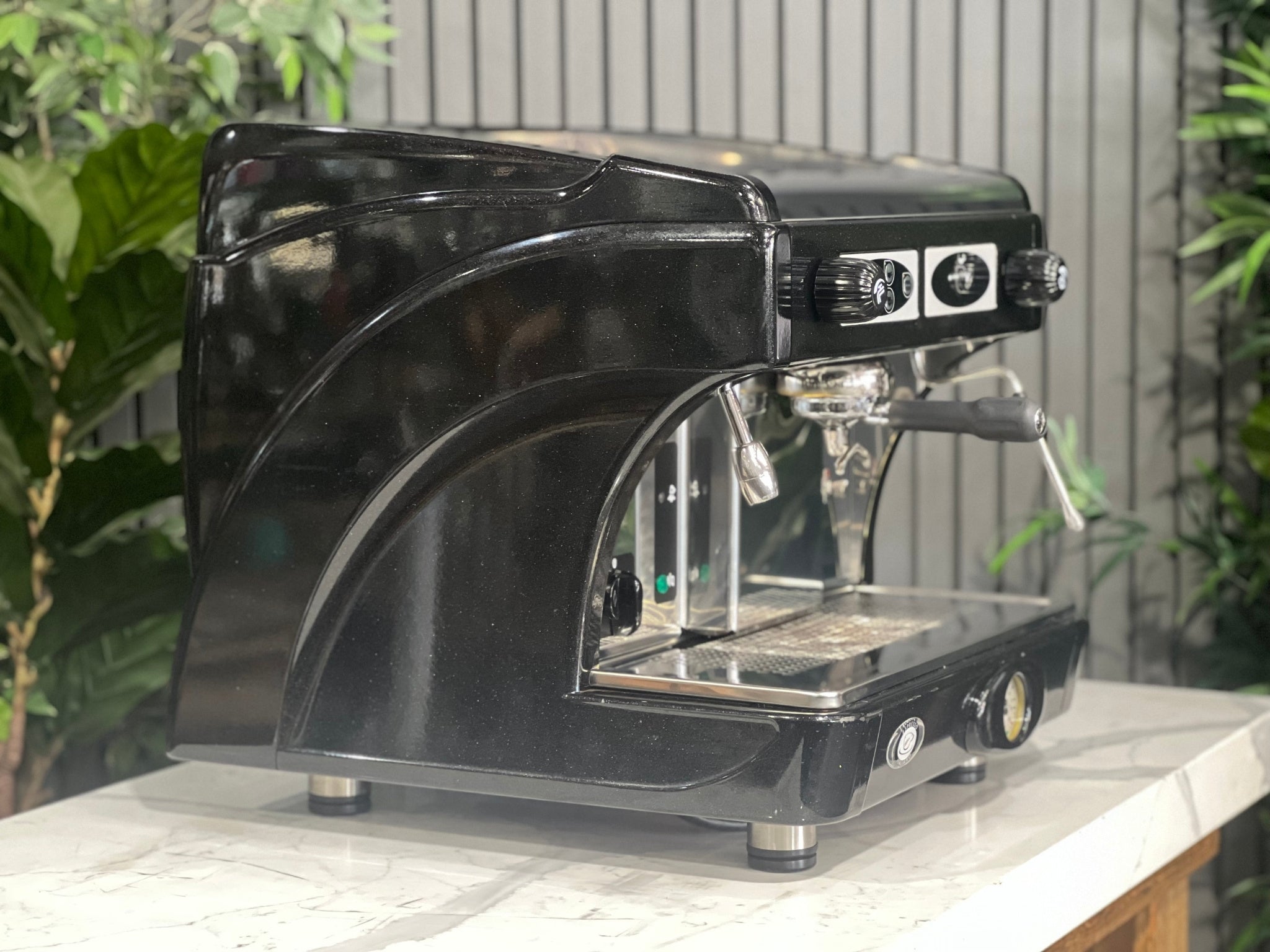 Astoria Calypso 1 Group Black Espresso Coffee Machine sitting on a marble bench with greenery in the background. Image is taken from an angle so that the  side of machine and part of the front is visible.