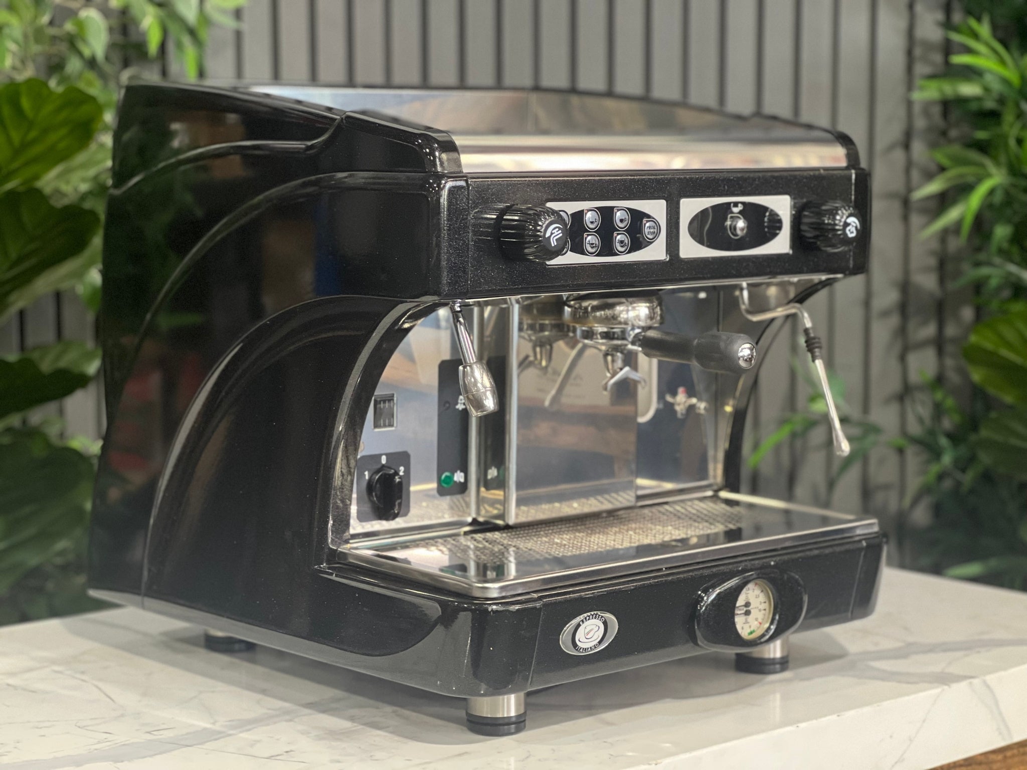 Astoria Calypso 1 Group Black Espresso Coffee Machine sitting on a marble bench with greenery in the background. Image is taken from an angle so that the front and side of machine is visible.