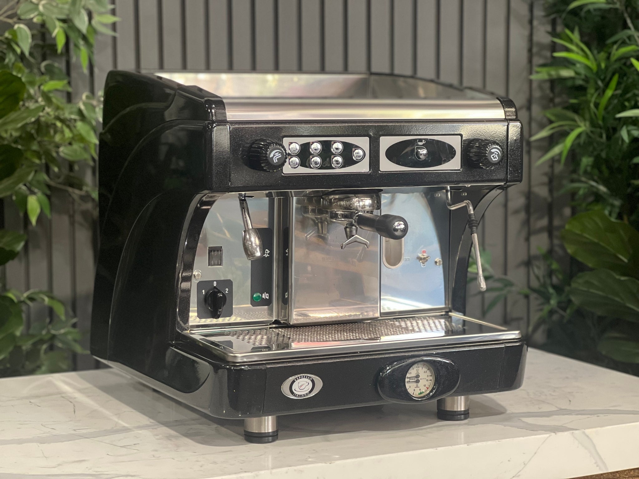 Astoria Calypso 1 Group Black Espresso Coffee Machine sitting on a marble bench with greenery in the background. Image is taken from an angle so that the front and side of machine is visible.