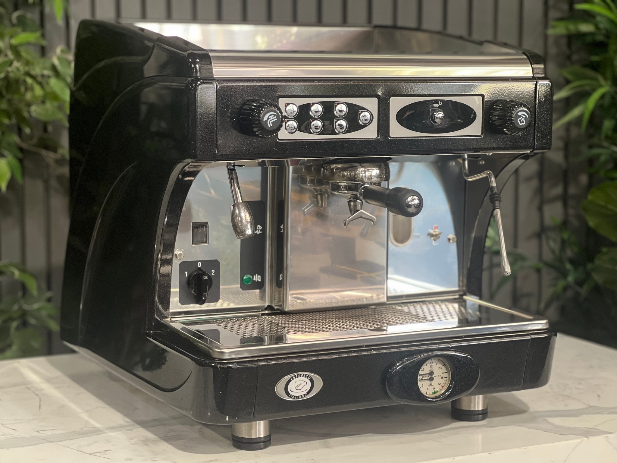 Astoria Calypso 1 Group Black Espresso Coffee Machine sitting on a marble bench with greenery in the background. Image is taken from an angle so that the front and side of machine is visible.