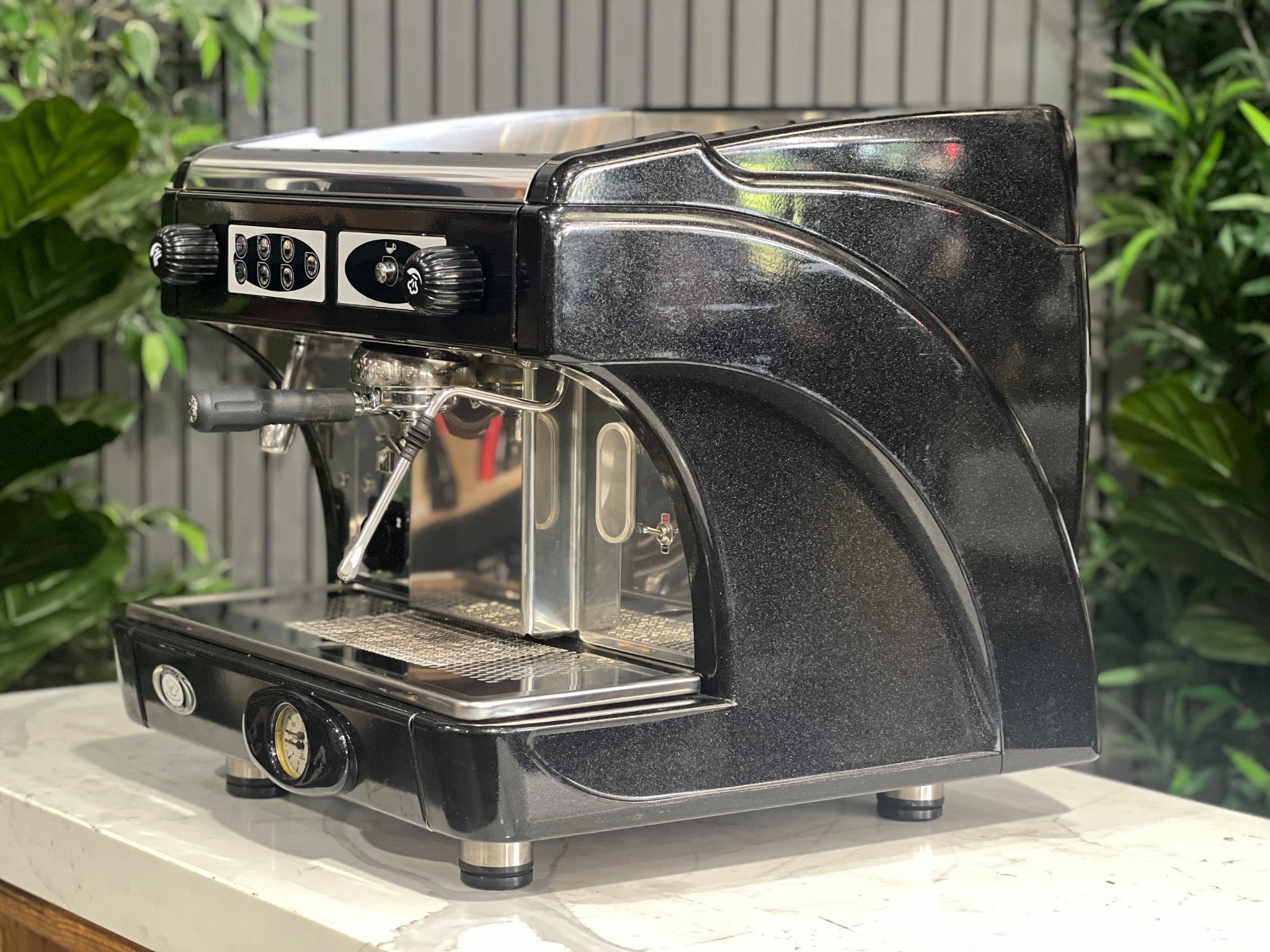 Astoria Calypso 1 Group Black Espresso Coffee Machine sitting on a marble bench with greenery in the background. Image is taken from an angle so that the front and side of machine is visible.