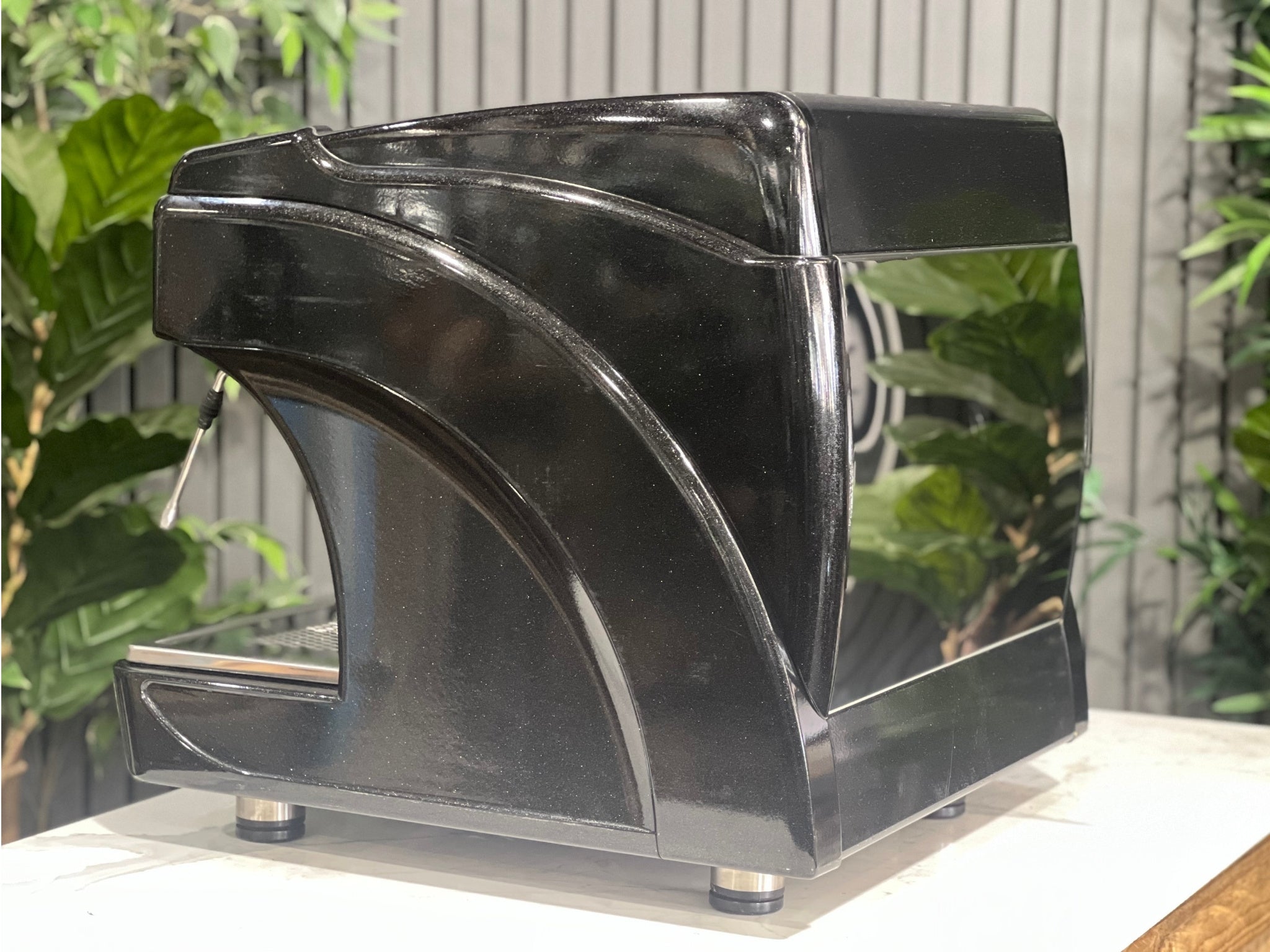 Astoria Calypso 1 Group Black Espresso Coffee Machine sitting on a marble bench with greenery in the background. Image is taken from an angle so that the back and side of machine is visible.