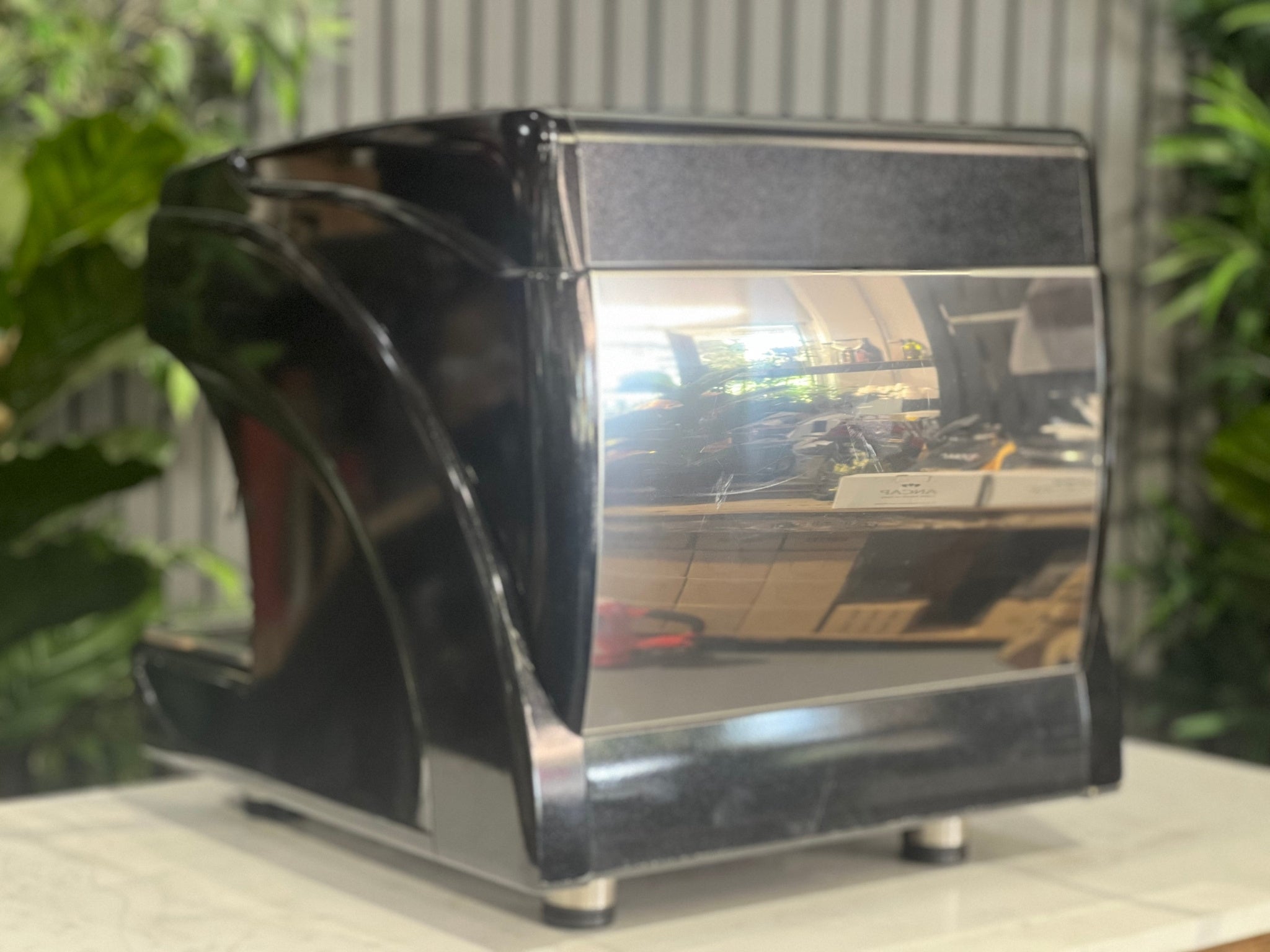Astoria Calypso 1 Group Black Espresso Coffee Machine sitting on a marble bench with greenery in the background. Image is taken from an angle behind the machine so that the back and side of machine is visible.