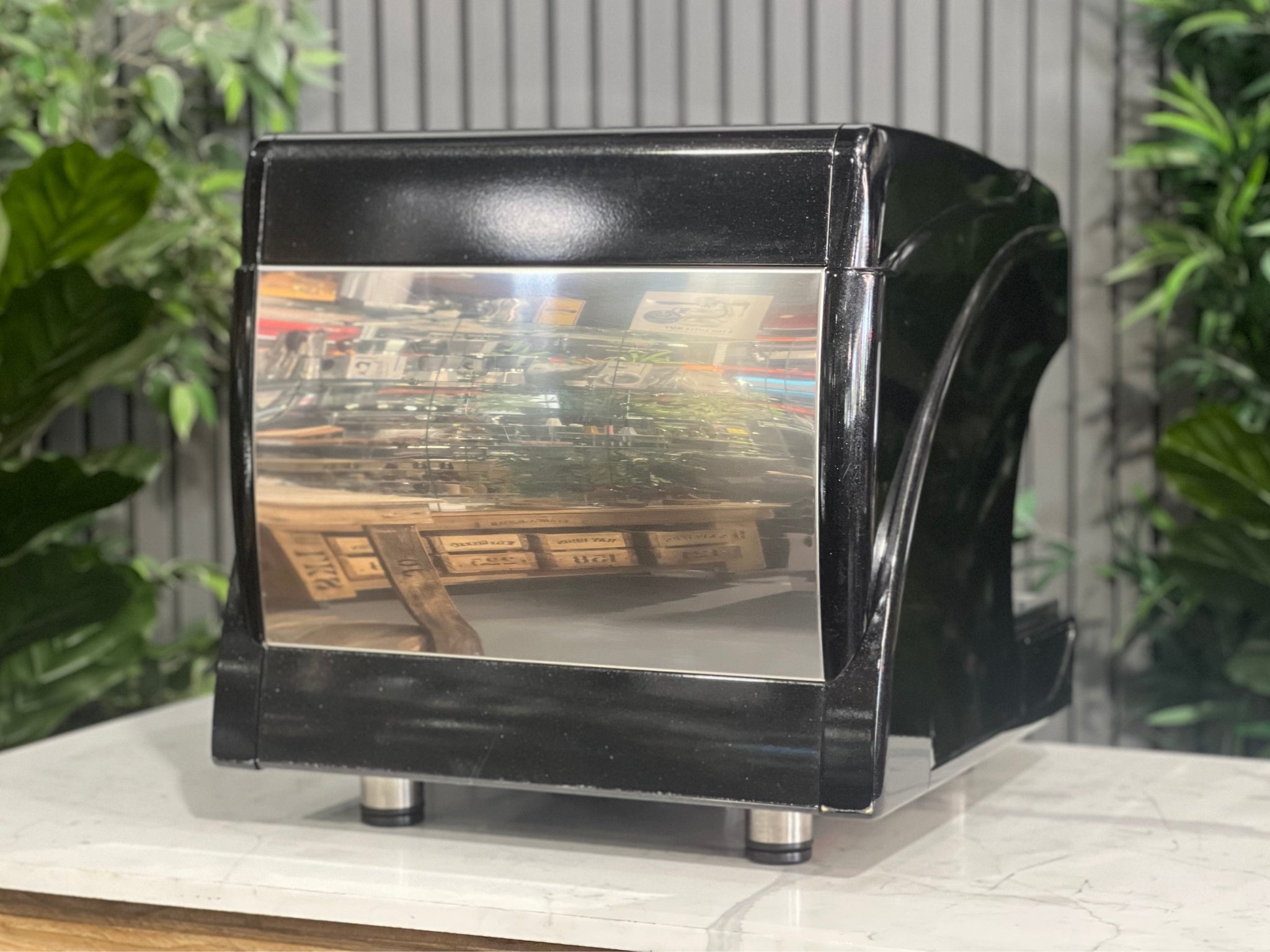 Astoria Calypso 1 Group Black Espresso Coffee Machine sitting on a marble bench with greenery in the background. Image is taken from behind the machine and on an angle so that the back and side of machine is visible.