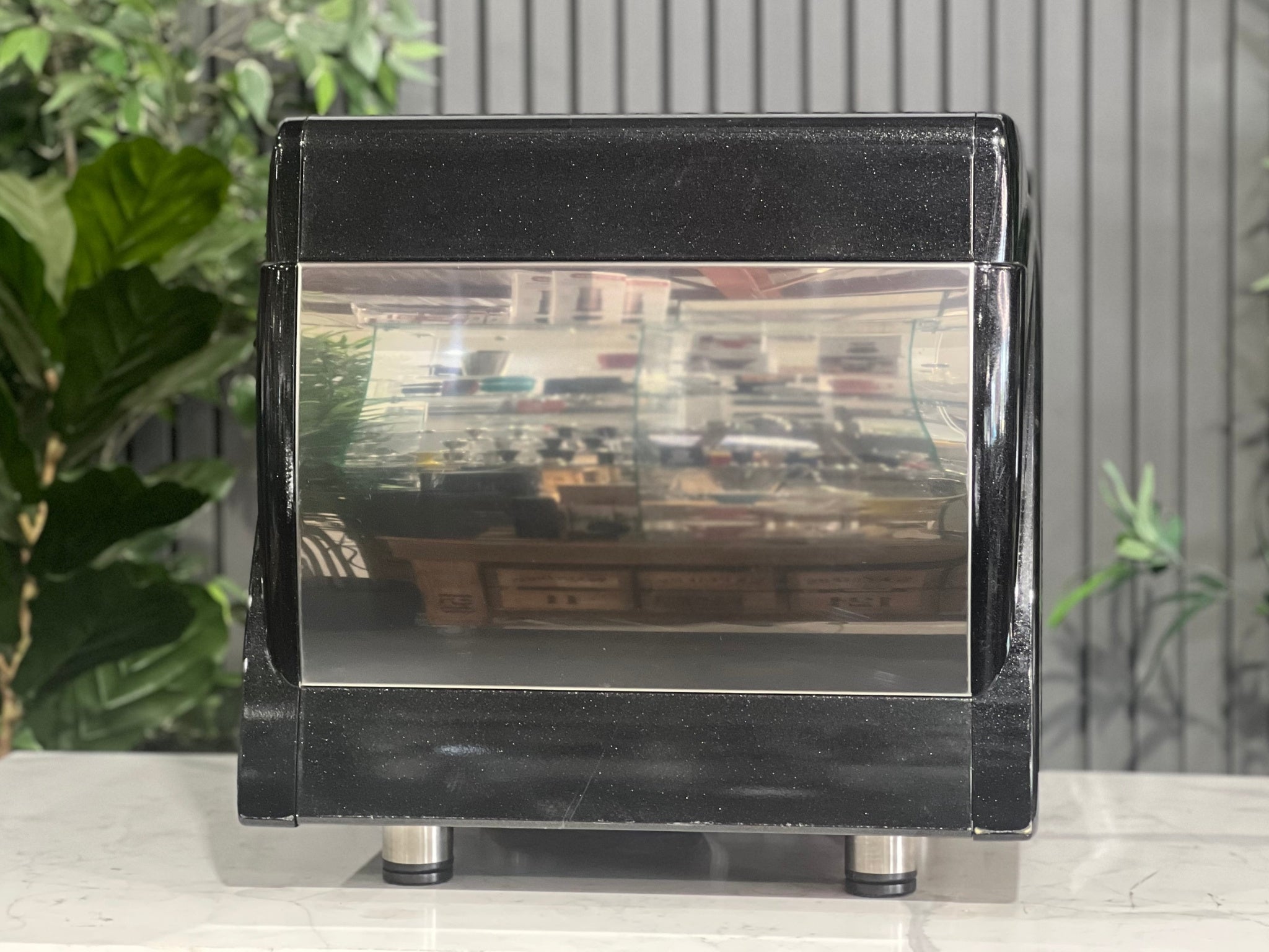 Astoria Calypso 1 Group Black Espresso Coffee Machine sitting on a marble bench with greenery in the background. Image is taken from behind the machine so the back of machine is visible.