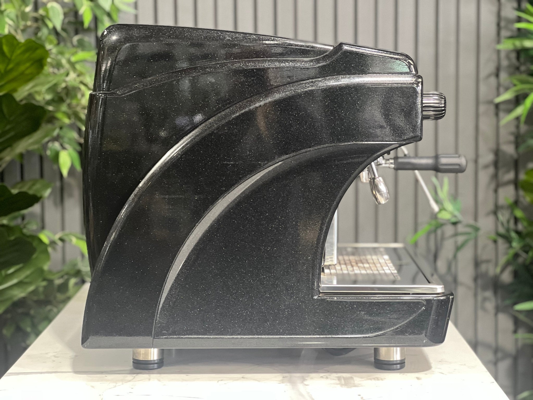 Astoria Calypso 1 Group Black Espresso Coffee Machine sitting on a marble bench with greenery in the background. Image is taken from the side of machine so the side of machine is visible.