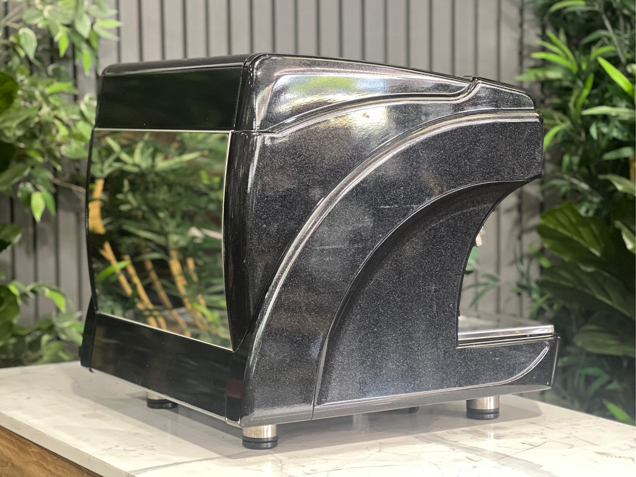 Astoria Calypso 1 Group Black Espresso Coffee Machine sitting on a marble bench with greenery in the background. Image is taken from an angle so that the back and side of machine is visible.