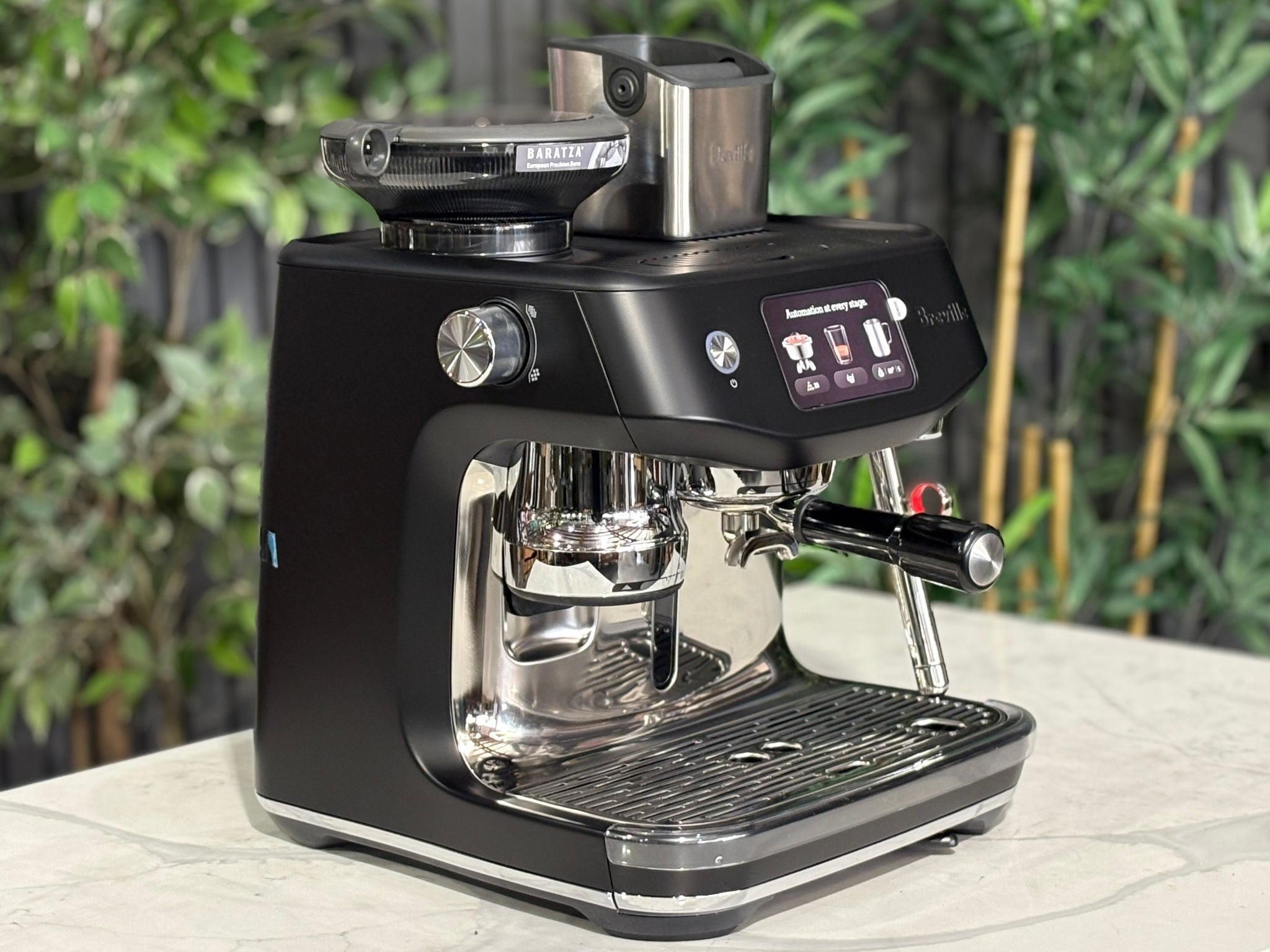 Breville Oracle Jet 1 Group Coffee Machine Black - New, angled view on a marble bench with greenery in the background, showing the black body, touch screen display, milk steamer, and hopper.