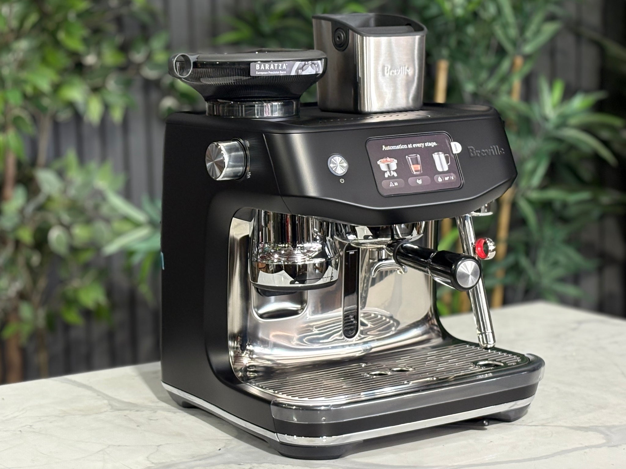 Breville Oracle Jet 1 Group Coffee Machine Black - New, angled view on a marble bench with greenery in the background, showing the black body, touch screen display, milk steamer, and hopper.