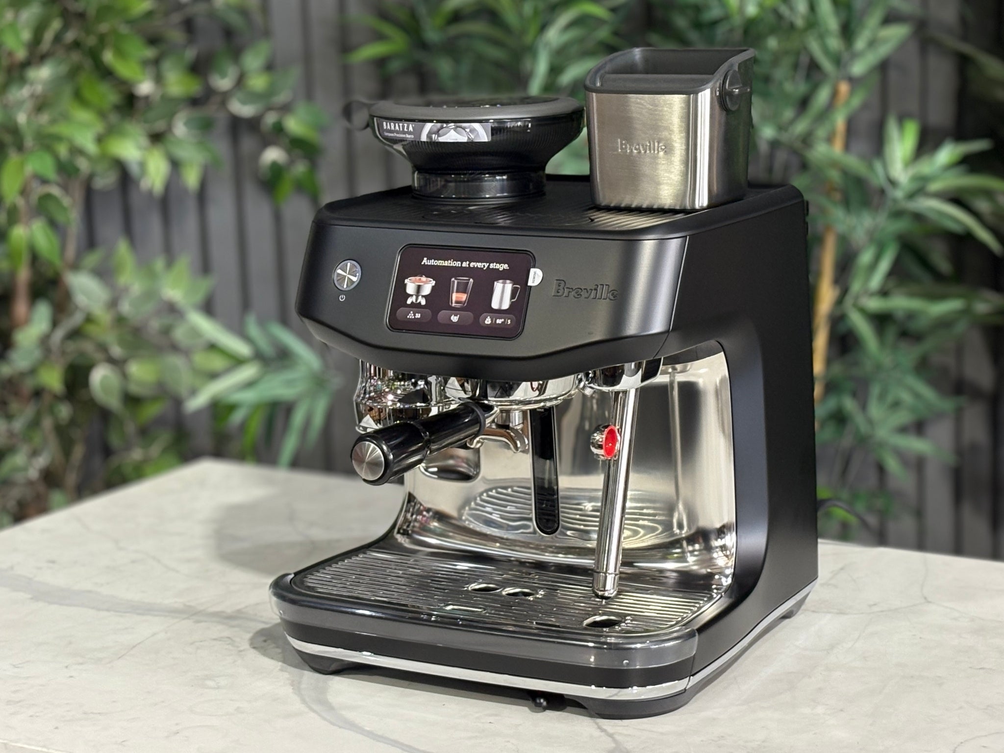 Breville Oracle Jet 1 Group Coffee Machine Black - New, angled view on a marble bench with greenery in the background, showing the black body, touch screen display, milk steamer, and hopper.