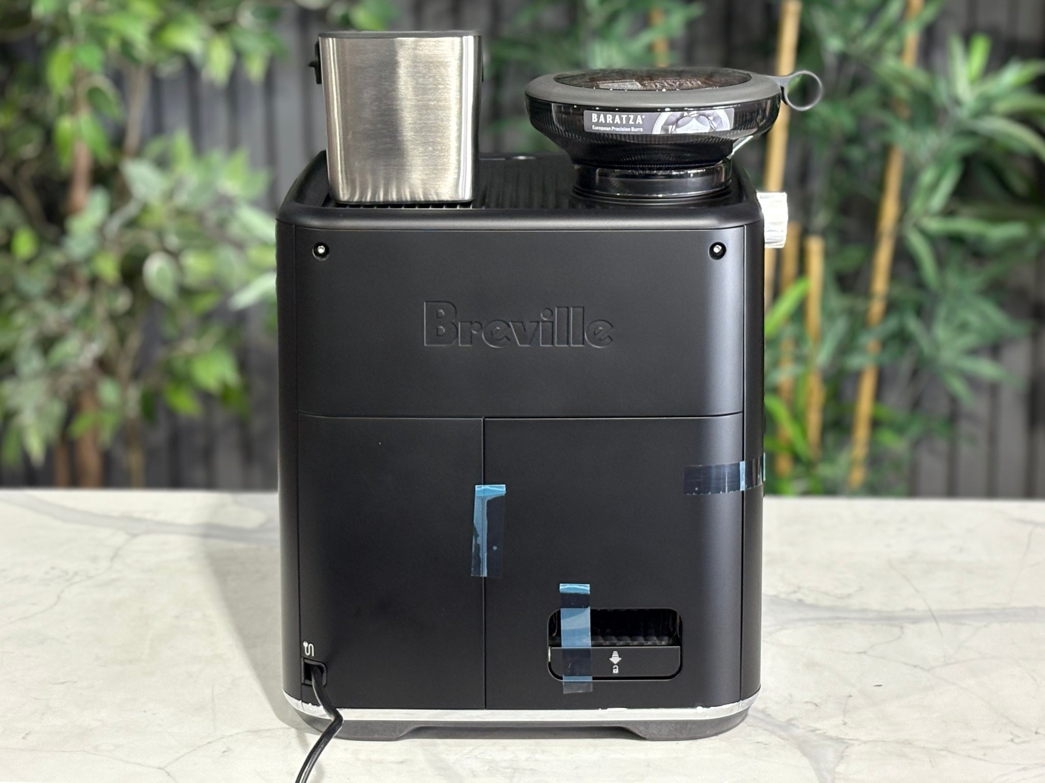 Breville Oracle Jet 1 Group Coffee Machine Black - New, rear view on a marble bench, showing the black body and power connection area with greenery in the background.