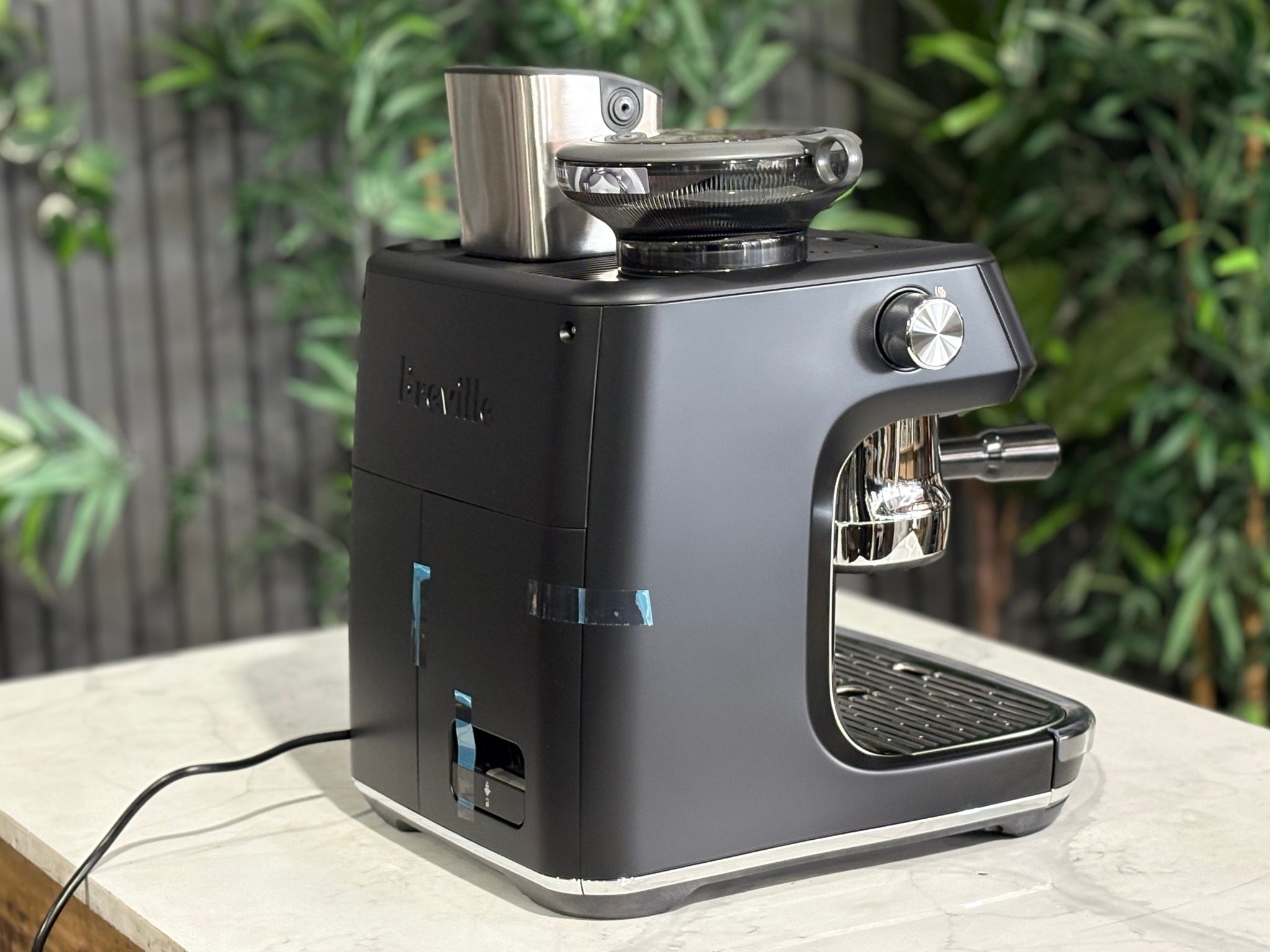 Breville Oracle Jet 1 Group Coffee Machine Black - New, angled rear view on a marble bench with greenery, showing the black body, water reservoir area, and overall sleek design.