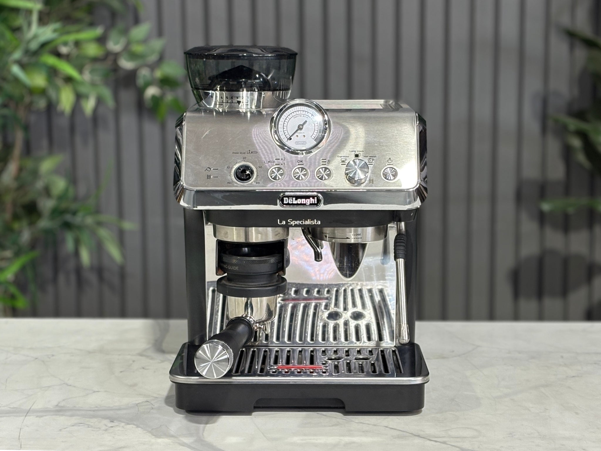 A Delonghi La Specialista Arte 1 Group Coffee Machine in Black sitting on a marble bench. There is greenery in the background. The image is taken from the front of the machine so that you can see the front of the machine, control panel, buttons and group head.