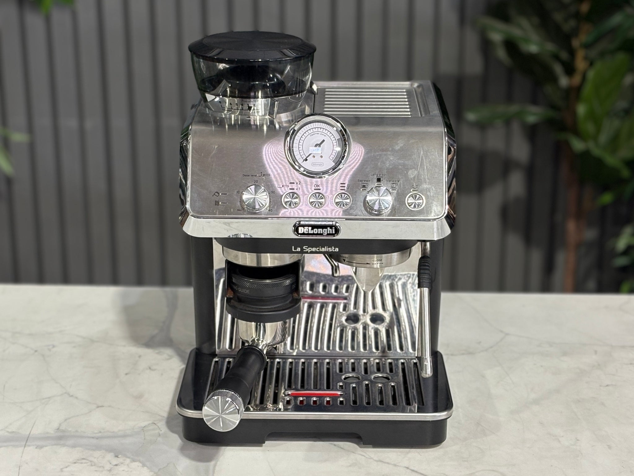 A Delonghi La Specialista Arte 1 Group Coffee Machine in Black sitting on a marble bench. There is greenery in the background. The photo is taken from a high front angle, clearly showcasing both the front panel and the top of the machine.