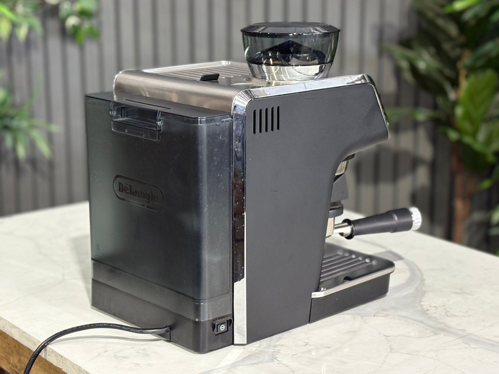 A Delonghi La Specialista Arte 1 Group Coffee Machine in Black sitting on a marble bench. There is greenery in the background. The image is taken from behind the machine and the machine is sitting on an angle so that you can see the back of the machine and the side.