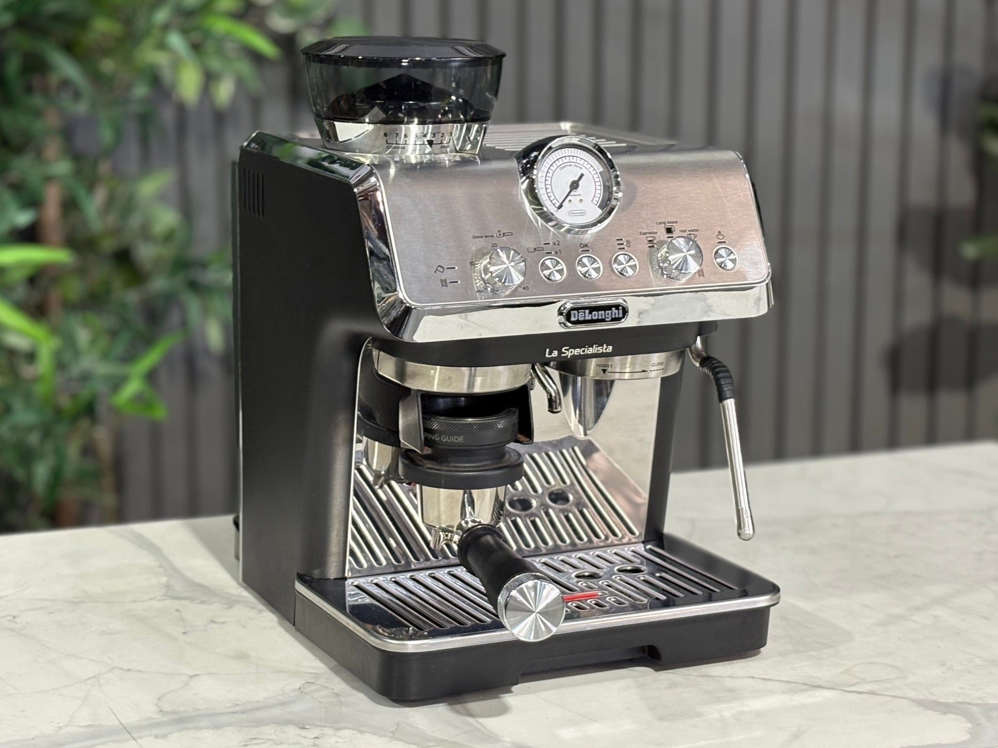 A Delonghi La Specialista Arte 1 Group Coffee Machine in Black is sitting on a marble bench. There is greenery in the background. The image is taken from the front of the machine and the machine is sitting on an angle so that you can see the front of the machine and the side.