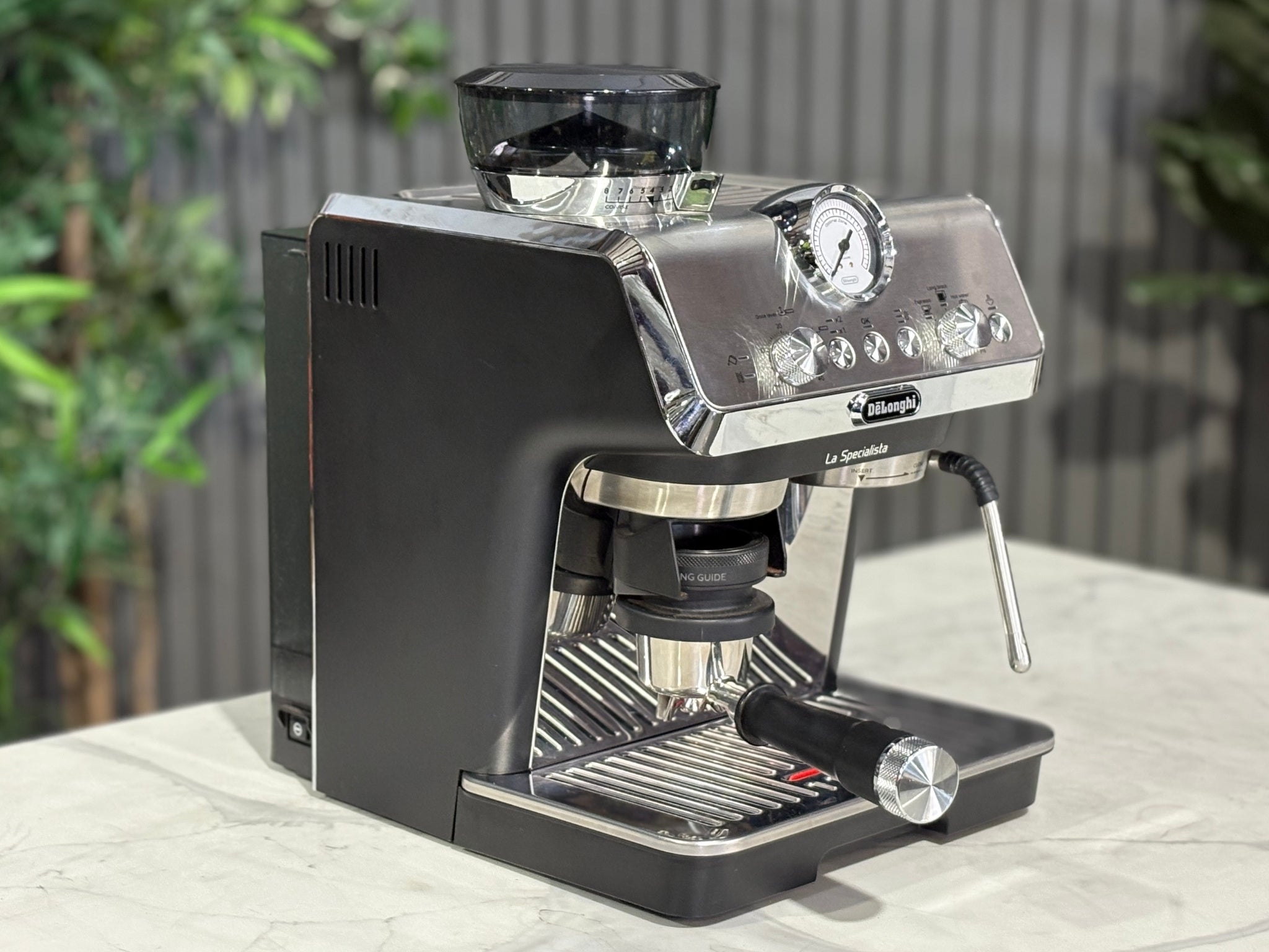 A Delonghi La Specialista Arte 1 Group Coffee Machine in Black is sitting on a marble bench. There is greenery in the background. The image is taken from the front of the machine and the machine is sitting on an angle so that you can see the front of the machine and the side.