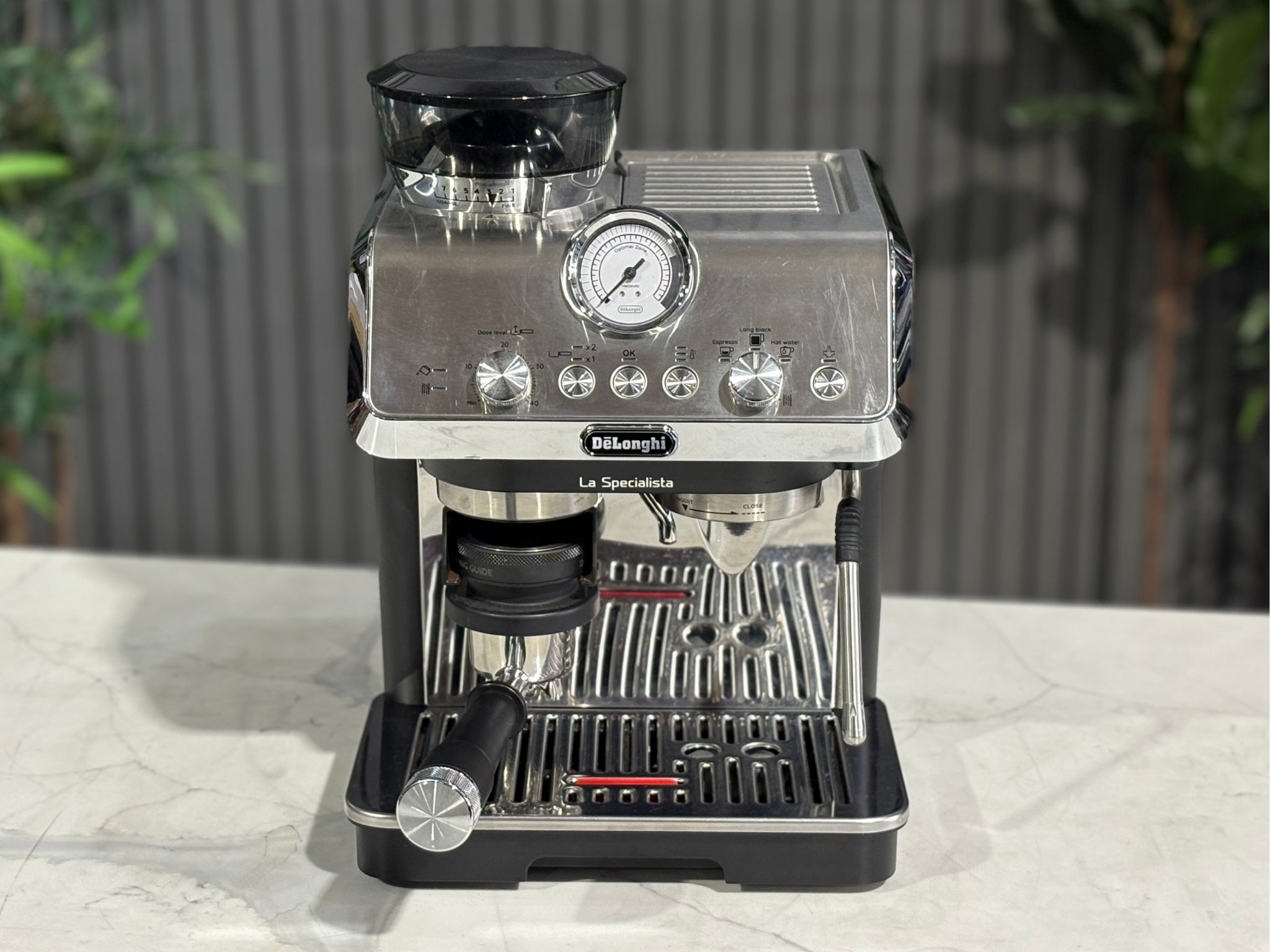 A Delonghi La Specialista Arte 1 Group Coffee Machine in Black sitting on a marble bench. There is greenery in the background. The photo is taken from a high front angle, clearly showcasing both the front panel and the top of the machine.