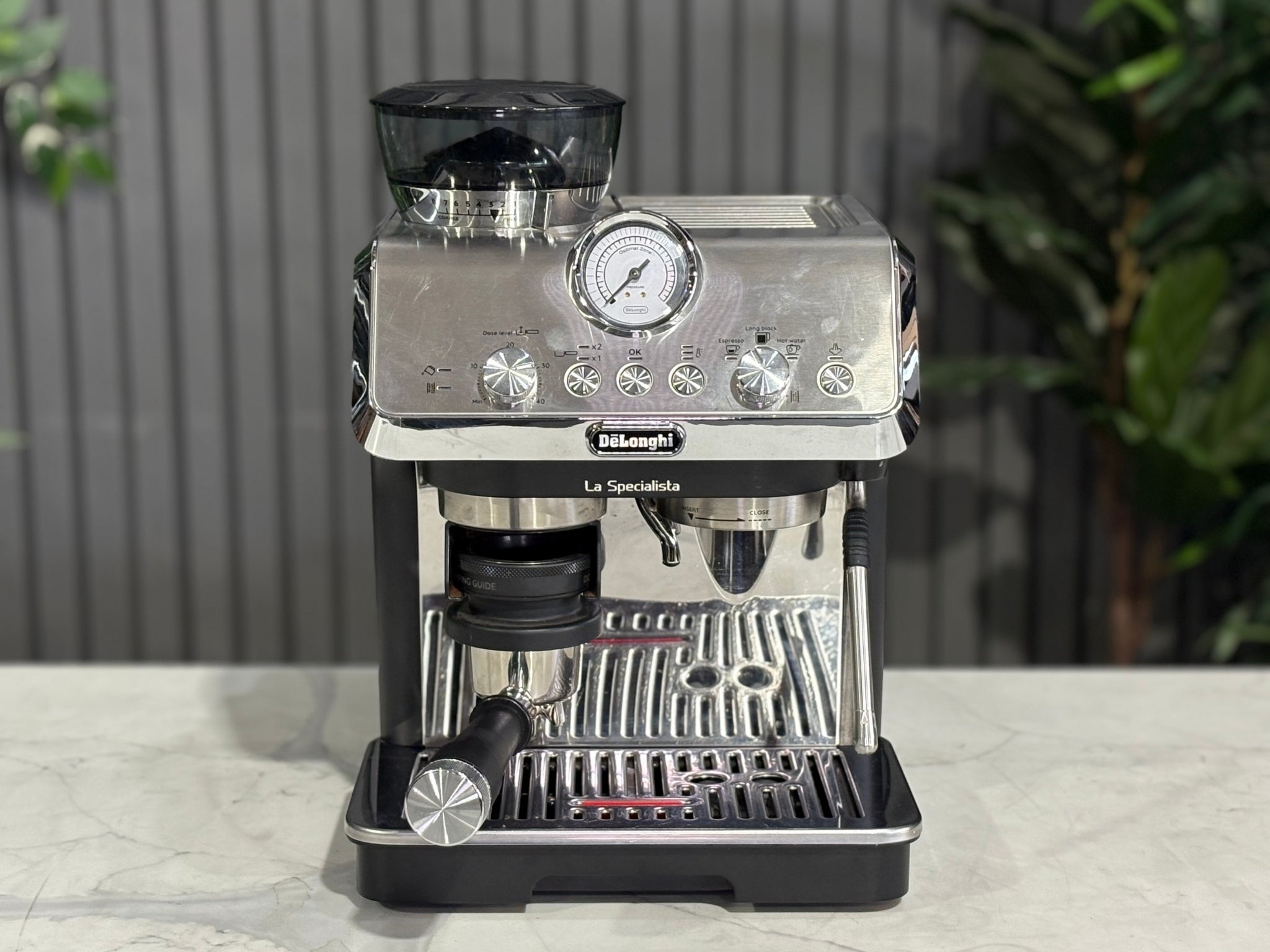 A Delonghi La Specialista Arte 1 Group Coffee Machine in Black sitting on a marble bench. There is greenery in the background. The image is taken from the front of the machine so that you can see the front of the machine, control panel, buttons and group head.