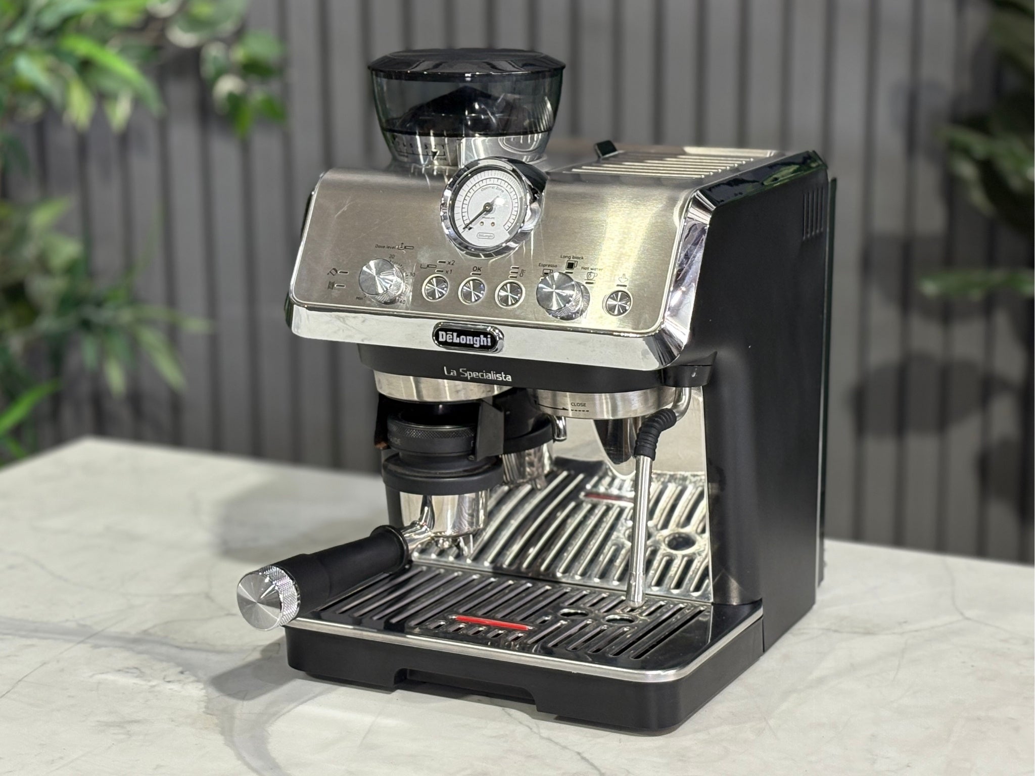 A Delonghi La Specialista Arte 1 Group Coffee Machine in Black is sitting on a marble bench. There is greenery in the background. The image is taken from the front of the machine and the machine is sitting on an angle so that you can see the front of the machine and the side.