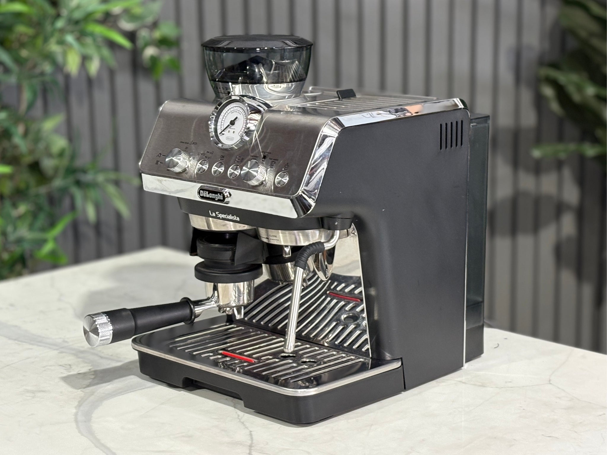 A Delonghi La Specialista Arte 1 Group Coffee Machine in Black is sitting on a marble bench. There is greenery in the background. The image is taken from the front of the machine and the machine is sitting on an angle so that you can see the front of the machine and the side.