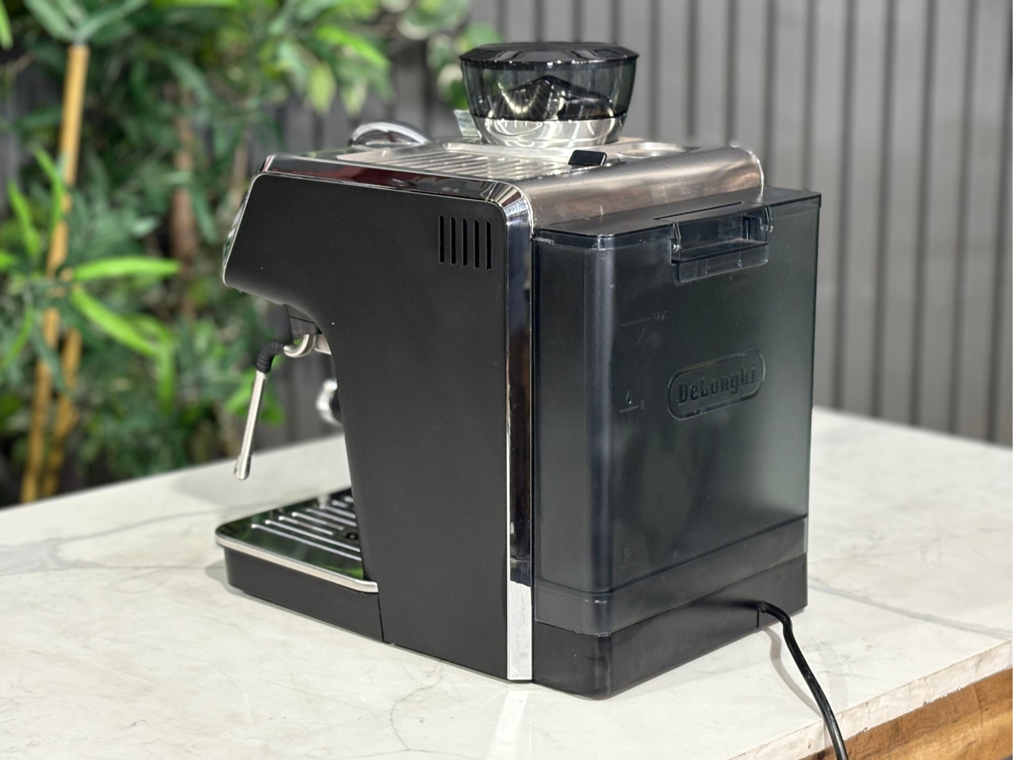 A Delonghi La Specialista Arte 1 Group Coffee Machine in Black sitting on a marble bench. There is greenery in the background. The image is taken from behind the machine and the machine is sitting on an angle so that you can see the back of the machine and the side.