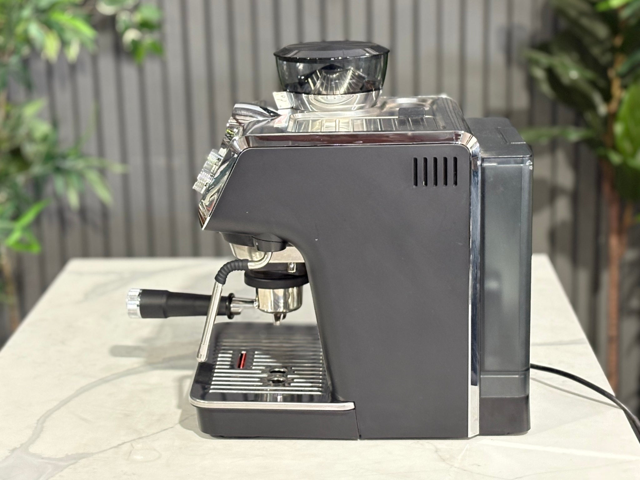 A Delonghi La Specialista Arte 1 Group Coffee Machine in Black sitting on a marble bench. There is greenery in the background. The image is taken from the side of the machine.