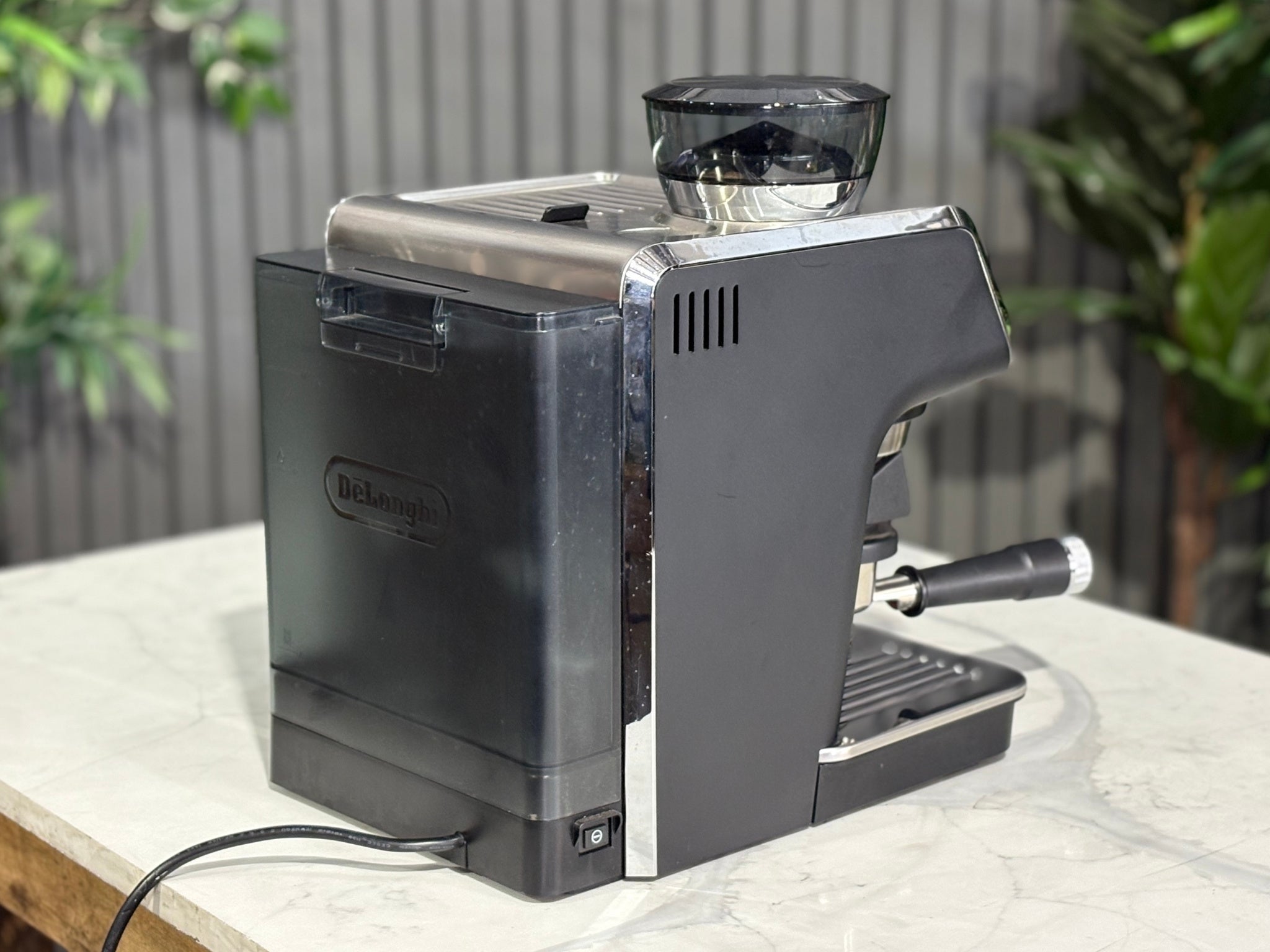 A Delonghi La Specialista Arte 1 Group Coffee Machine in Black sitting on a marble bench. There is greenery in the background. The image is taken from behind the machine and the machine is sitting on an angle so that you can see the back of the machine and the side.