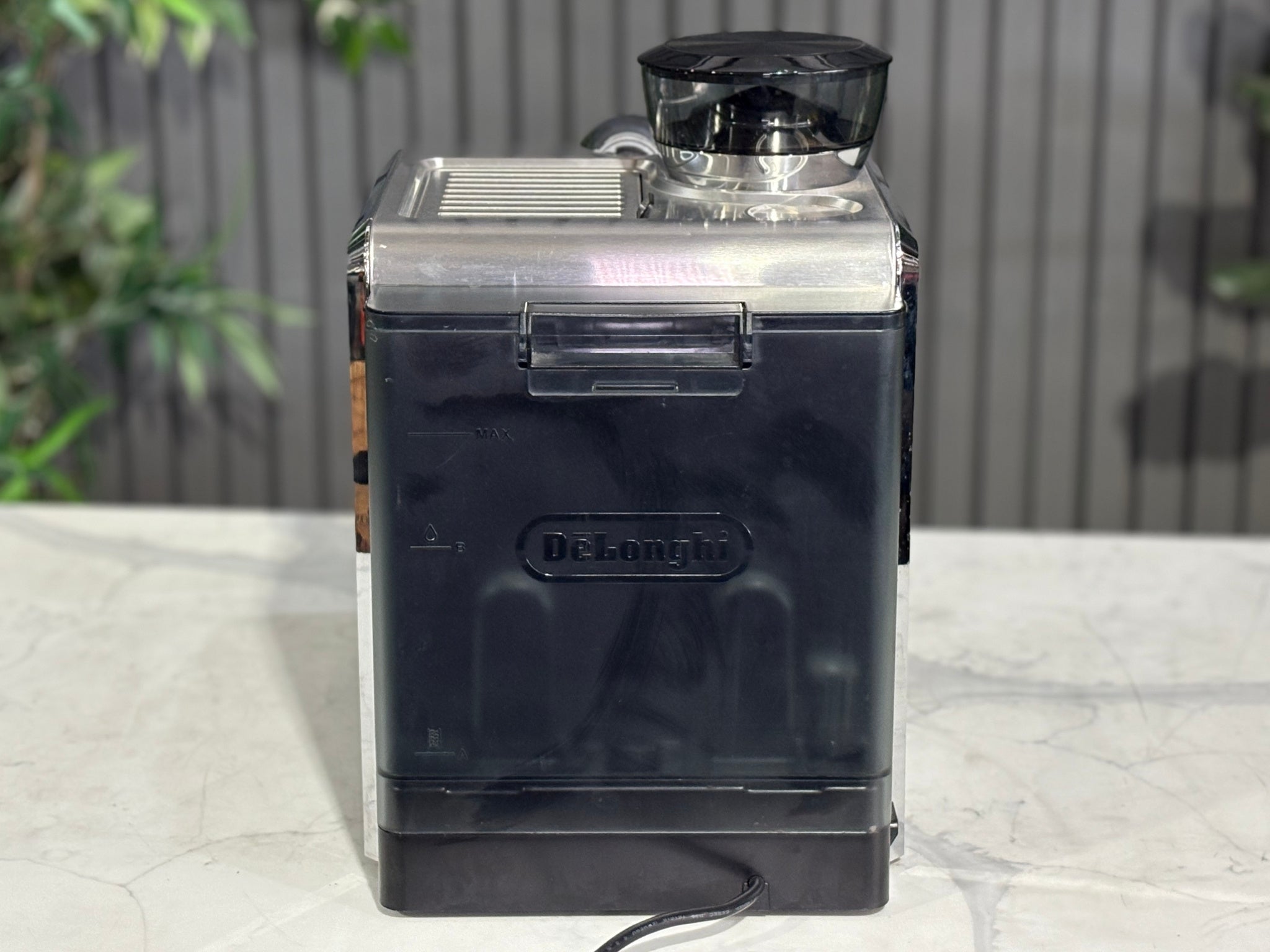 A Delonghi La Specialista Arte 1 Group Coffee Machine in Black is sitting on a marble bench. There is greenery in the background. The image is taken from behind the machine so the back is visible.